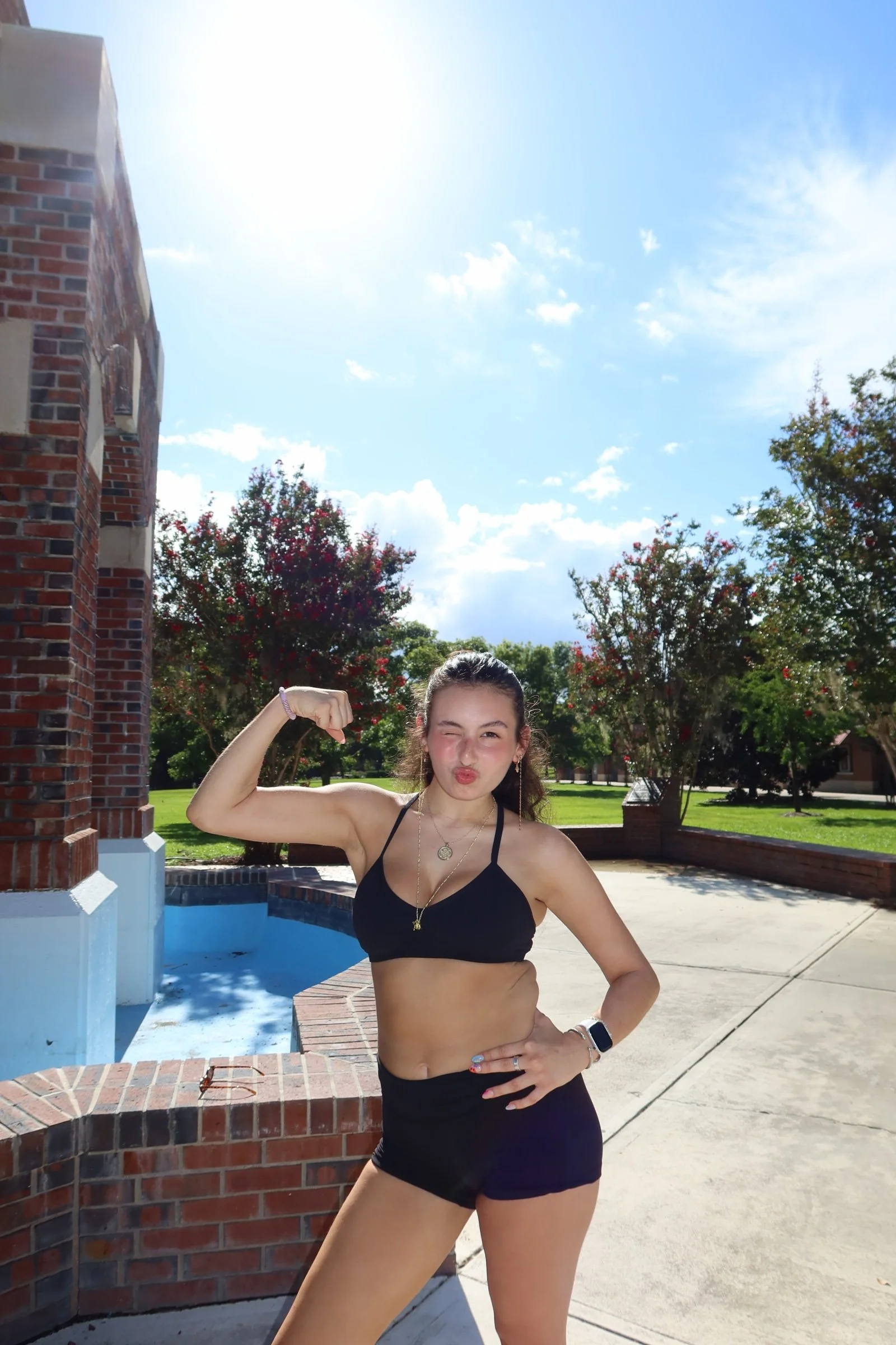 Young woman in a black sports bra and shorts striking a flex pose outdoors during daytime.