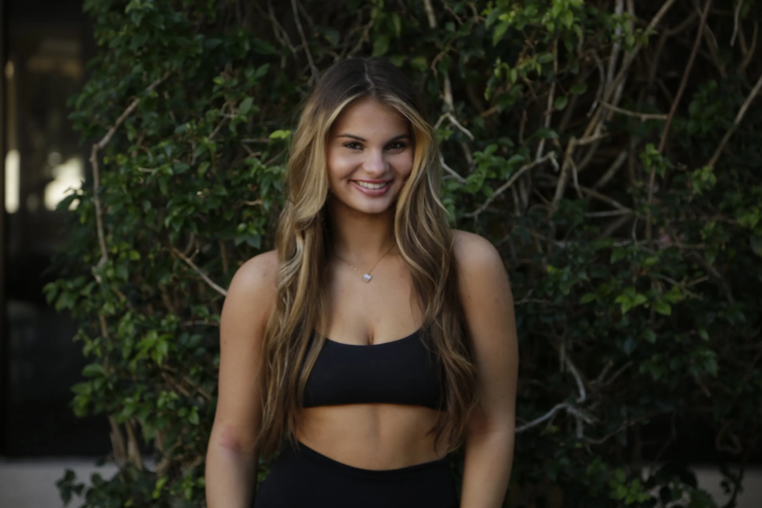 A young woman with long, wavy, light brown hair and light skin, smiling, standing outdoors in front of a green leafy background, wearing a black sports bra and a necklace with a small charm.