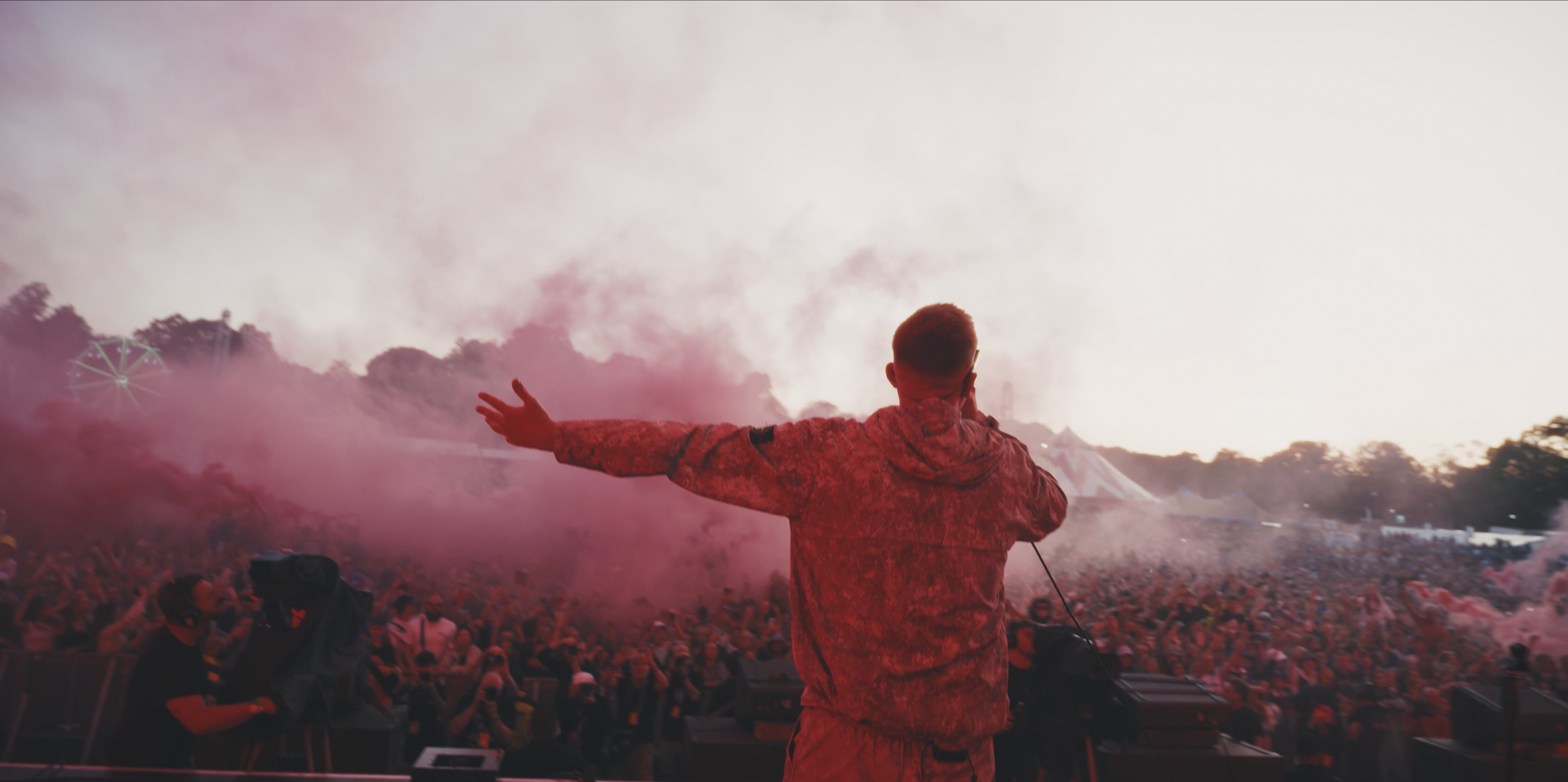 The Reytons Headline Tramlines 2025 Performer on stage with back to camera, smoking red powder, facing large crowd at outdoor music festival during sunset.