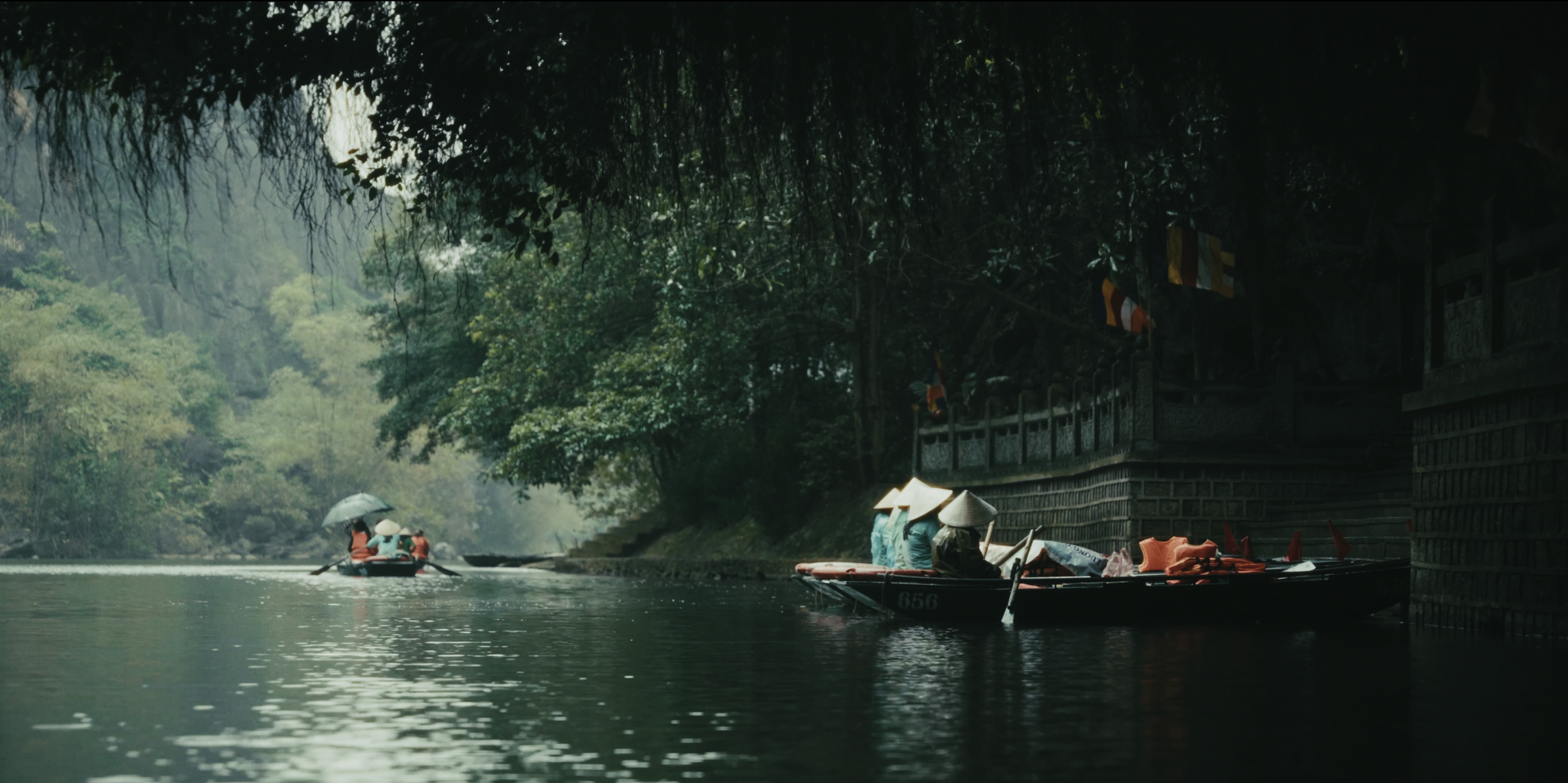 Boats on a river with lush green trees overhead and a stone wall along the riverbank in Vietnam.