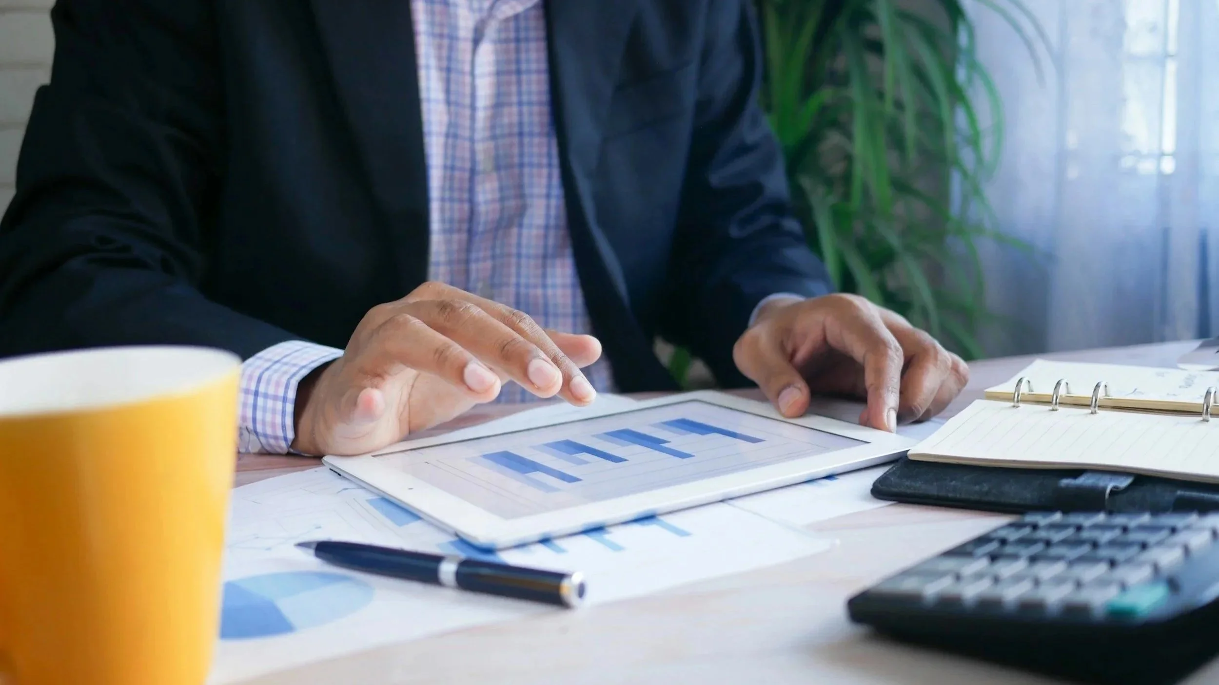 A person in a business suit using a tablet displaying a bar graph, with office supplies on a desk including a yellow mug, pen, notebook, and calculator, with a plant and window in the background.