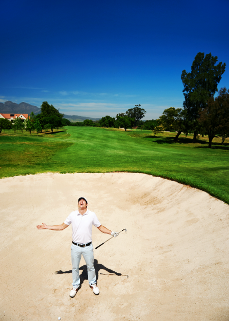 A man standing in a sand trap on a golf course with arms outstretched and holding a golf club, with a lush green fairway and trees in the background under a clear blue sky.