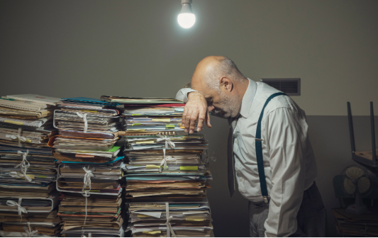 A man in a white shirt and suspenders leaning over a large pile of disorganized papers in an office, under a single overhead light.