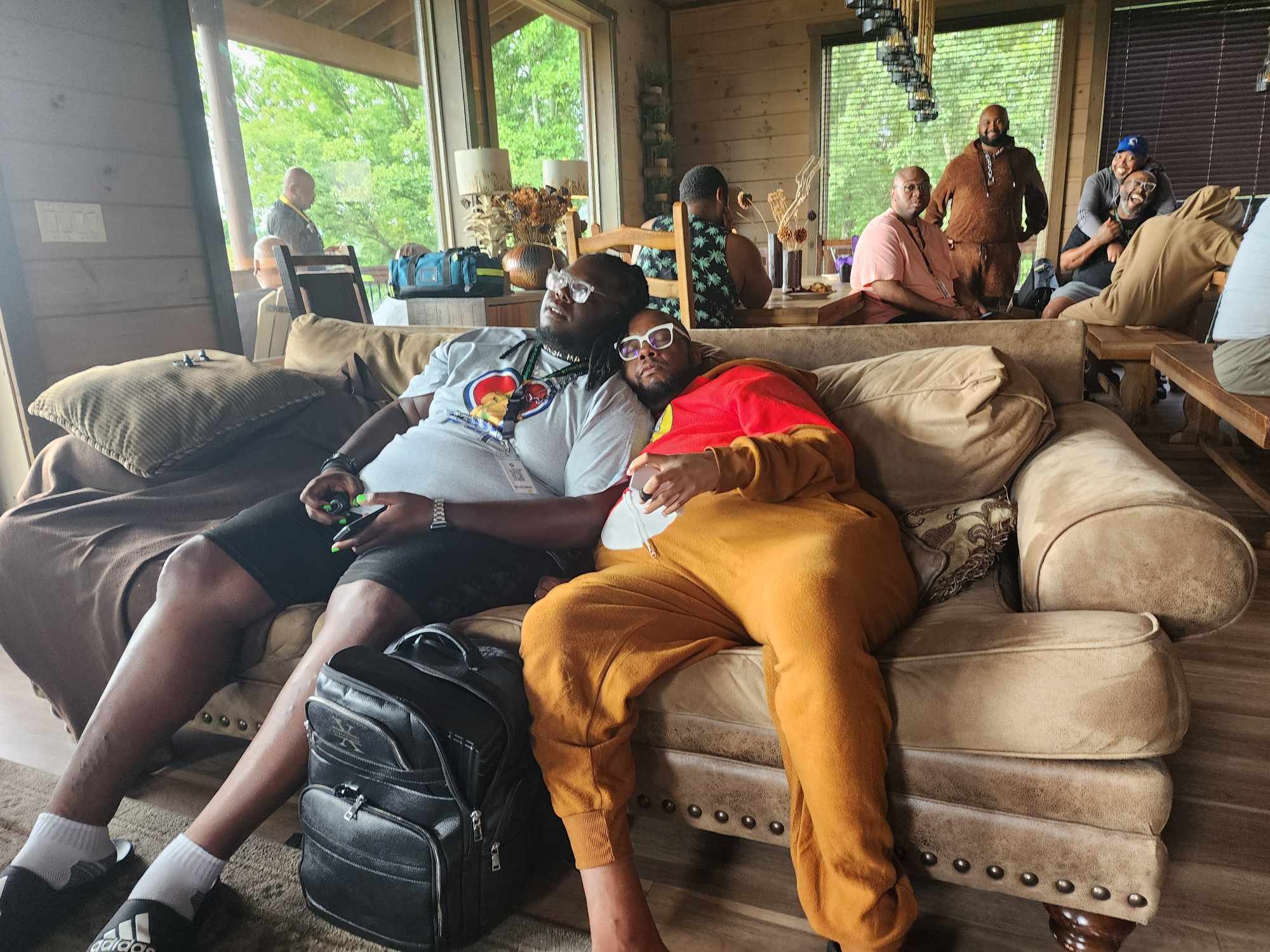 Group of people socializing and relaxing in a cozy living room with large windows showing greenery outside. Two individuals are lying on a couch, one resting their head on the other's lap. Several other people sit and stand around the room, engaging 