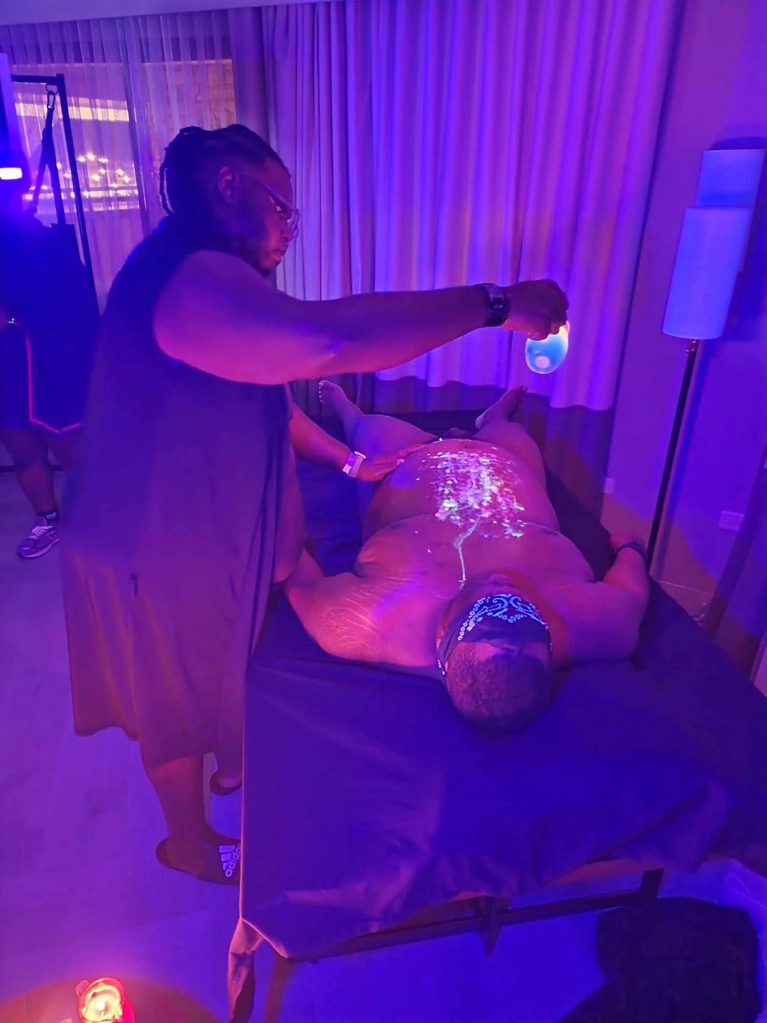 Man lying face down on massage table under blue and purple lights, receiving a treatment from a practitioner holding a glowing ball over the man's back in a dimly lit room.