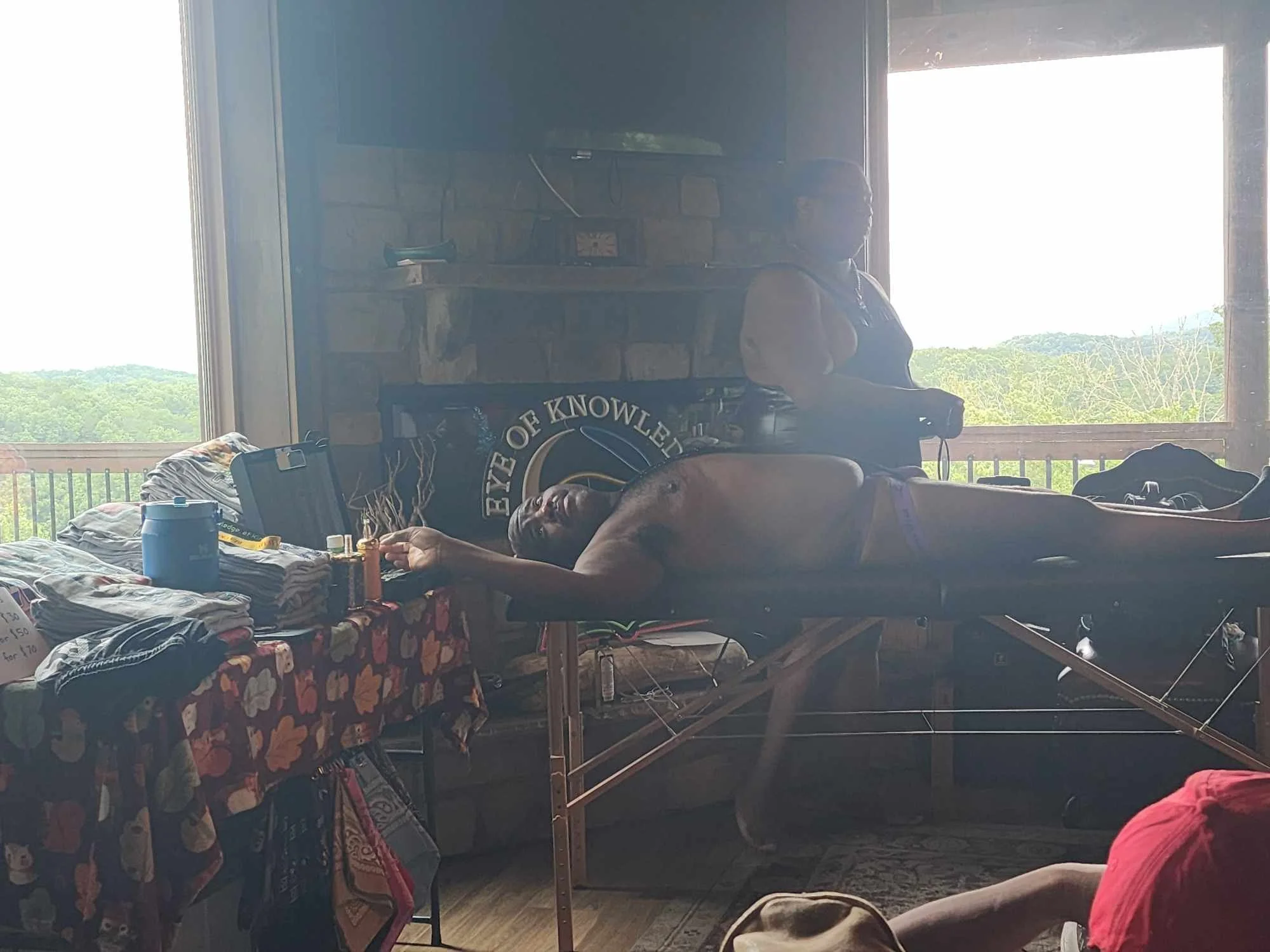 A person lying down on a bed with a collection of folded clothes on a table beside them, in a room with large windows showing a scenic view of green trees and hills, and a brick fireplace in the background.