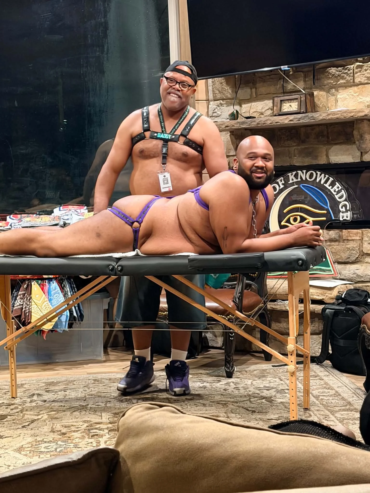 Two men in a living room dressed in BDSM gear, engaging in a consensual BDSM scene. One person, with glasses and a hat, is standing and smiling, while the other person, lying on a padded table, is nude and wearing a collar. The background features a 