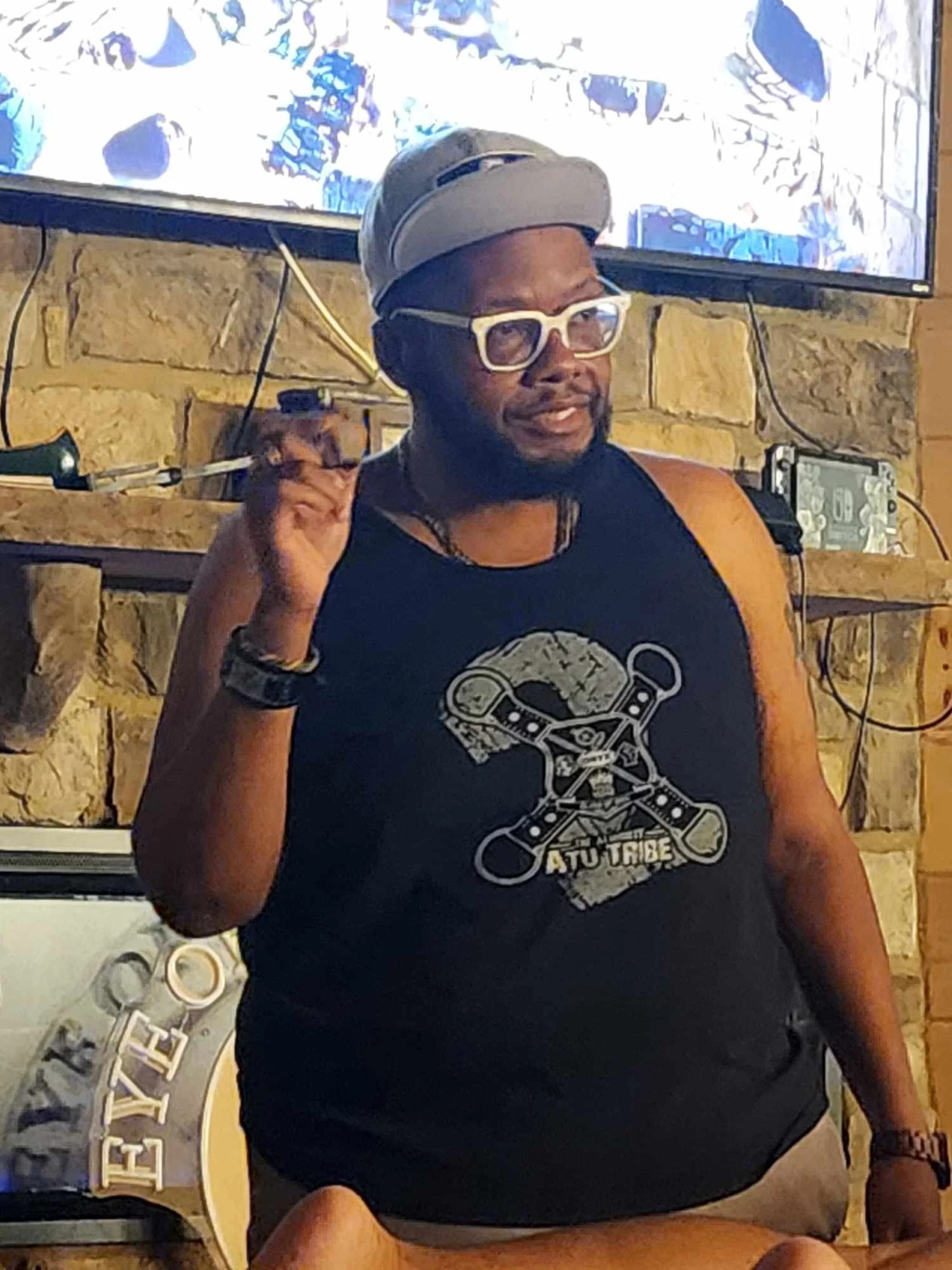 A man with glasses, a cap, and a black tank top sitting indoors with a television and a stone wall behind him, gesturing with his right hand.