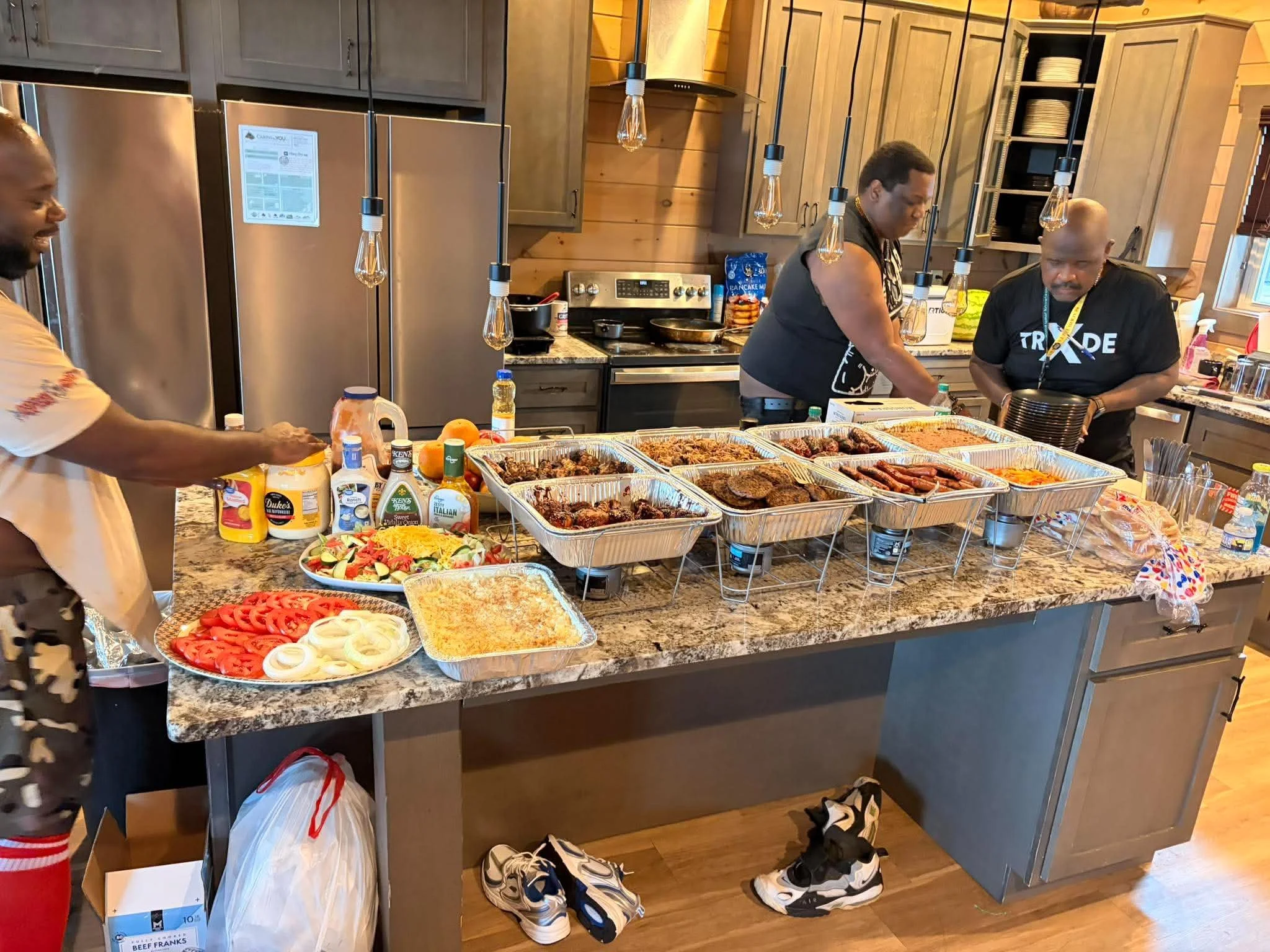 A kitchen countertop filled with various dishes including grilled meats, sliced vegetables, and salads. In the background, three people are preparing and serving food. The kitchen has wooden cabinets, hanging light bulbs, and a stainless steel refrig