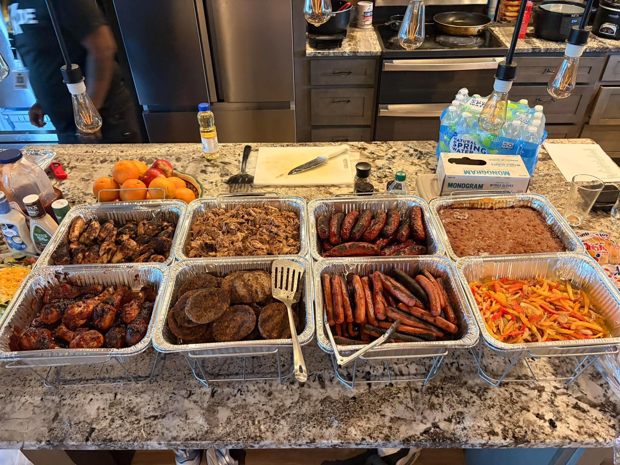 A kitchen counter with aluminum trays filled with grilled meats, sausages, and cooked vegetables. A white cutting board with a knife, bottles of water, and other condiments are on the counter, with a bundle of bottled water on the right side.