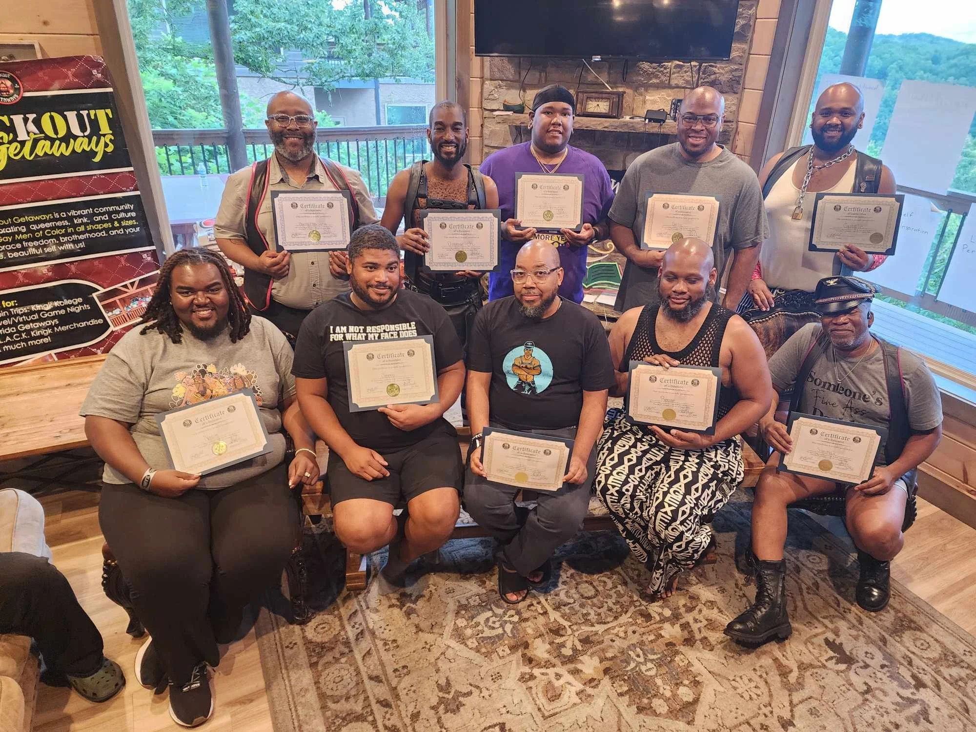 A diverse group of people in a cozy room holding certificates, posing for a group photo during an award or graduation ceremony. There are adults of different ages and styles, with some seated and others standing, smiling at the camera. The room featu