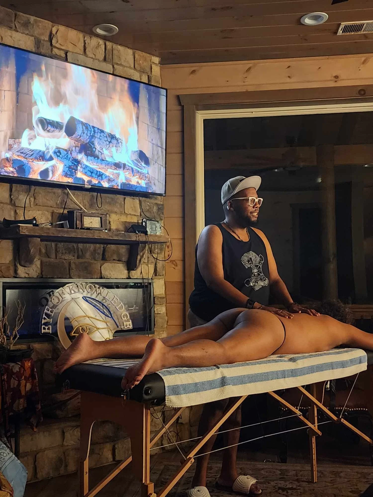 A person lying face down on a massage table during a massage, with a massage therapist standing beside them, in a room with a stone wall and a television showing a fireplace scene.