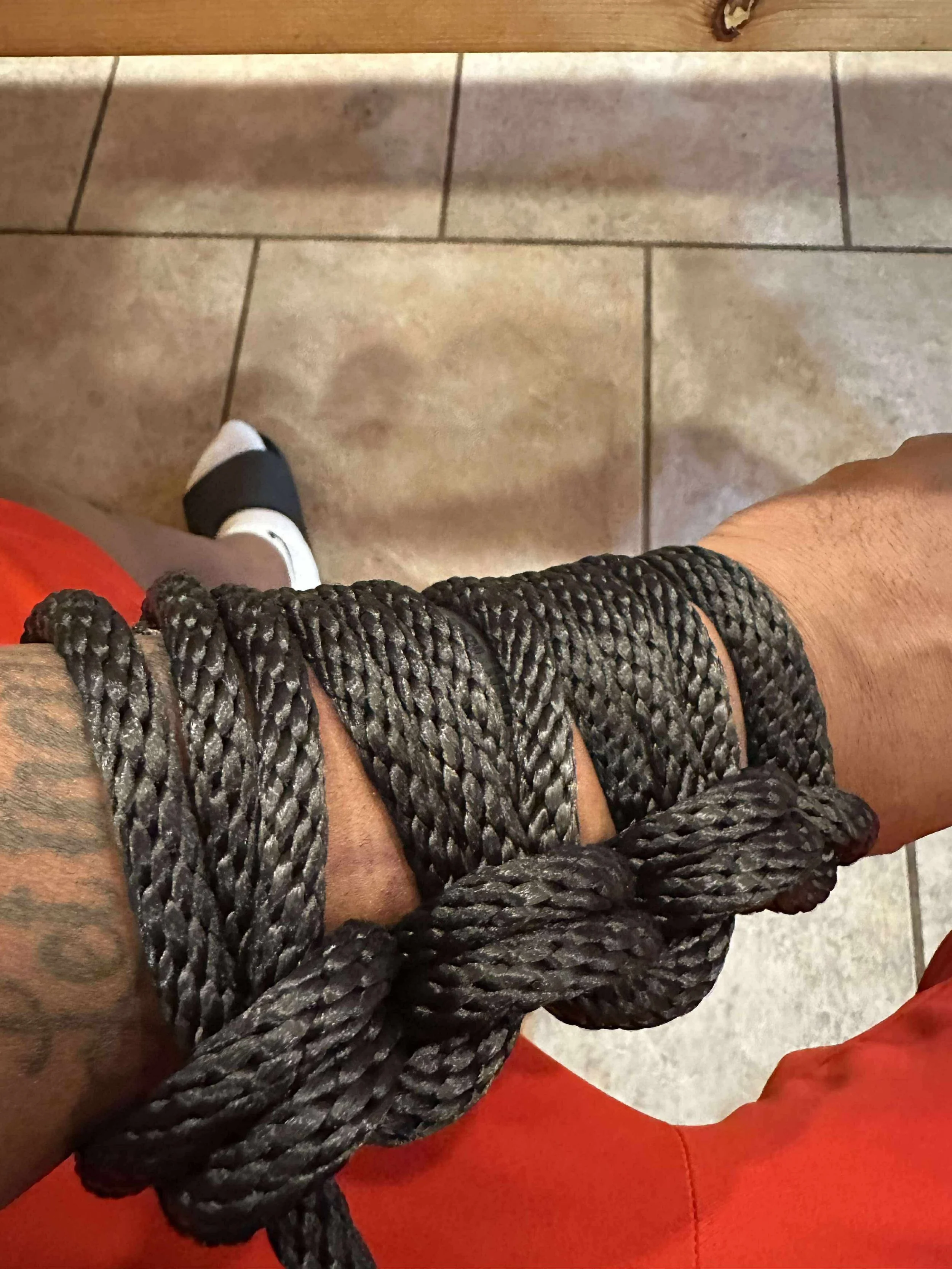 A person with a tattoo on their arm wearing a black rope band, a black and white shoe, and orange pants, standing on beige tiled floor next to a wooden surface.