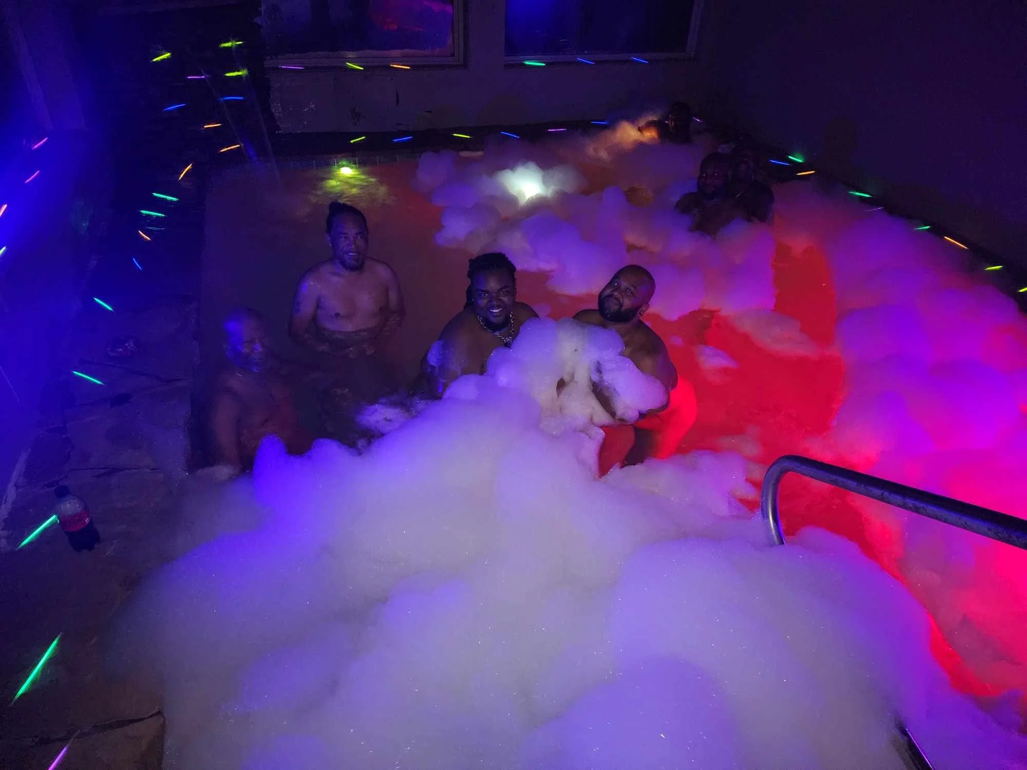 Four men enjoying themselves in a bubble-filled, purple and blue neon-lit indoor hot tub or pool.