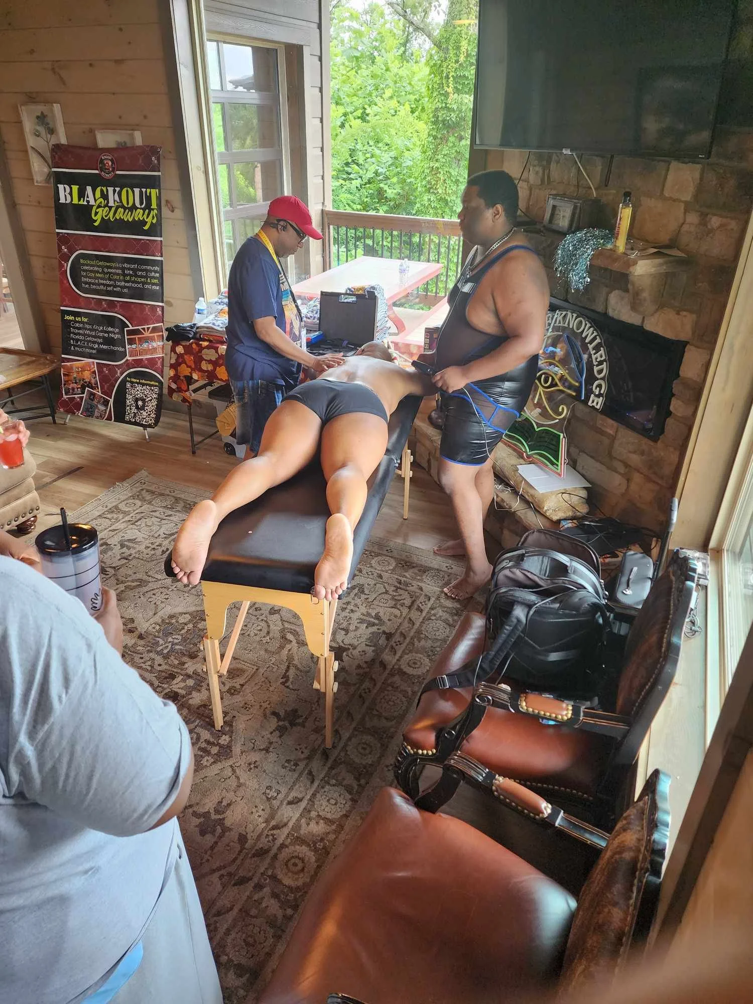 A person lying face down on a massage table receiving a massage from a man in black shorts. The setting appears to be a massage or wellness room with natural light, a TV mounted on the wall, and a banner that reads "Black Out Getaways".