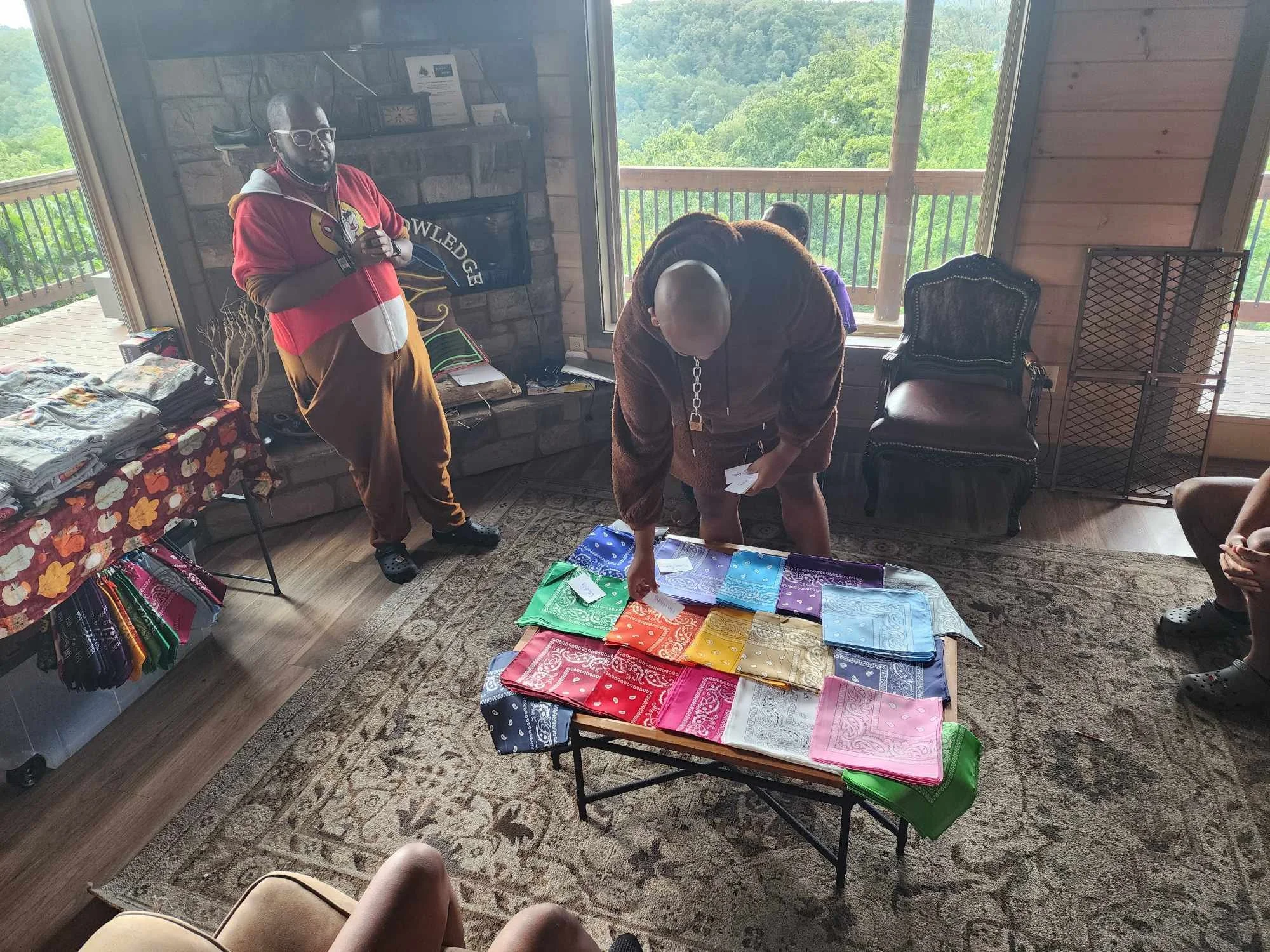 Three people are inside a room with wooden walls and large windows overlooking a green, forested landscape. A table is covered with multicolored bandanas, similar to handkerchiefs, arranged in stacks. One person is standing near the fireplace, wearin