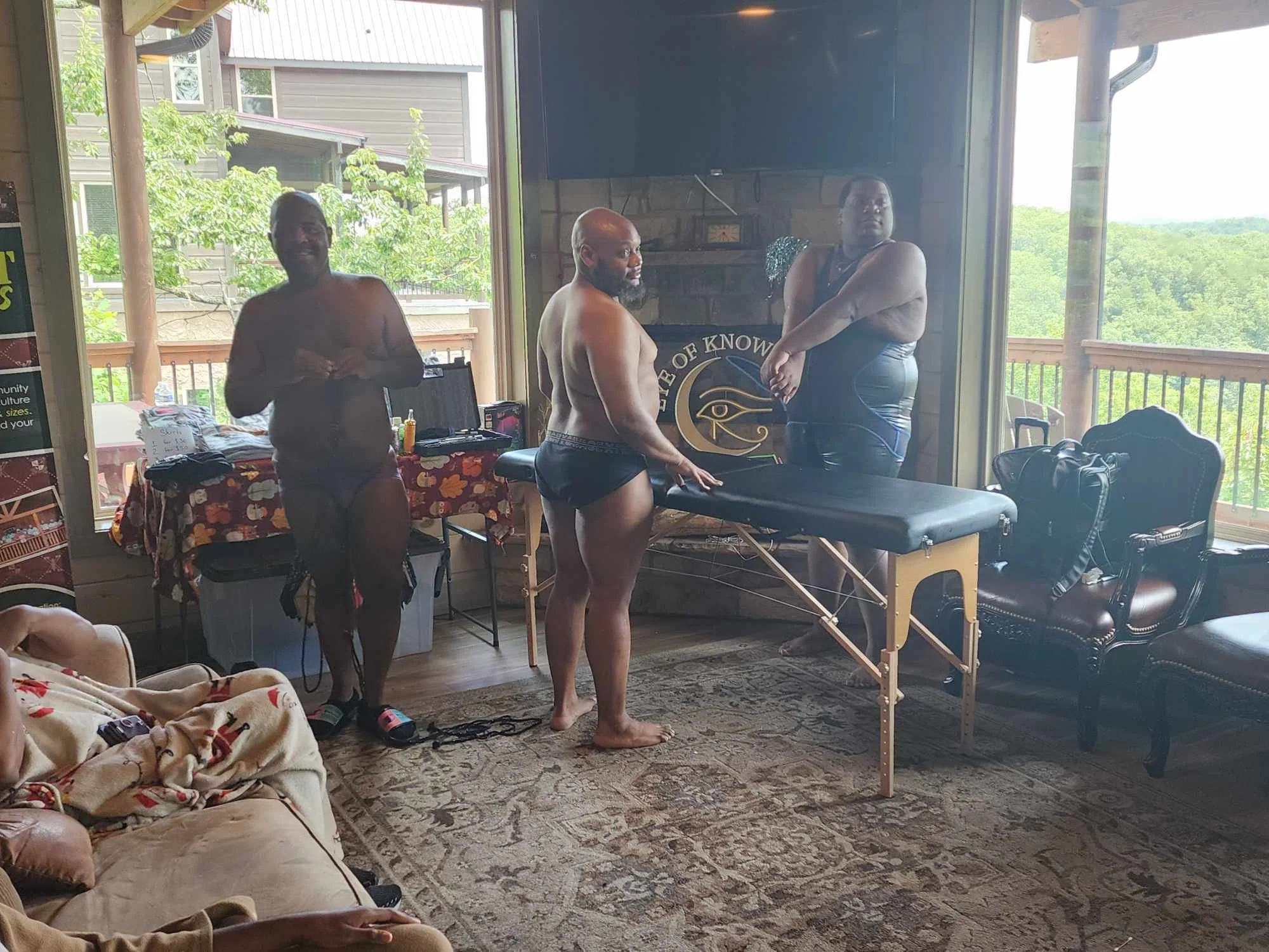 Three shirtless men in underwear standing around a massage table inside a room with large windows, with one man seated on the table and the other two standing nearby. There is a sign on a table with items for sale, and a logo on the wall that says 'T