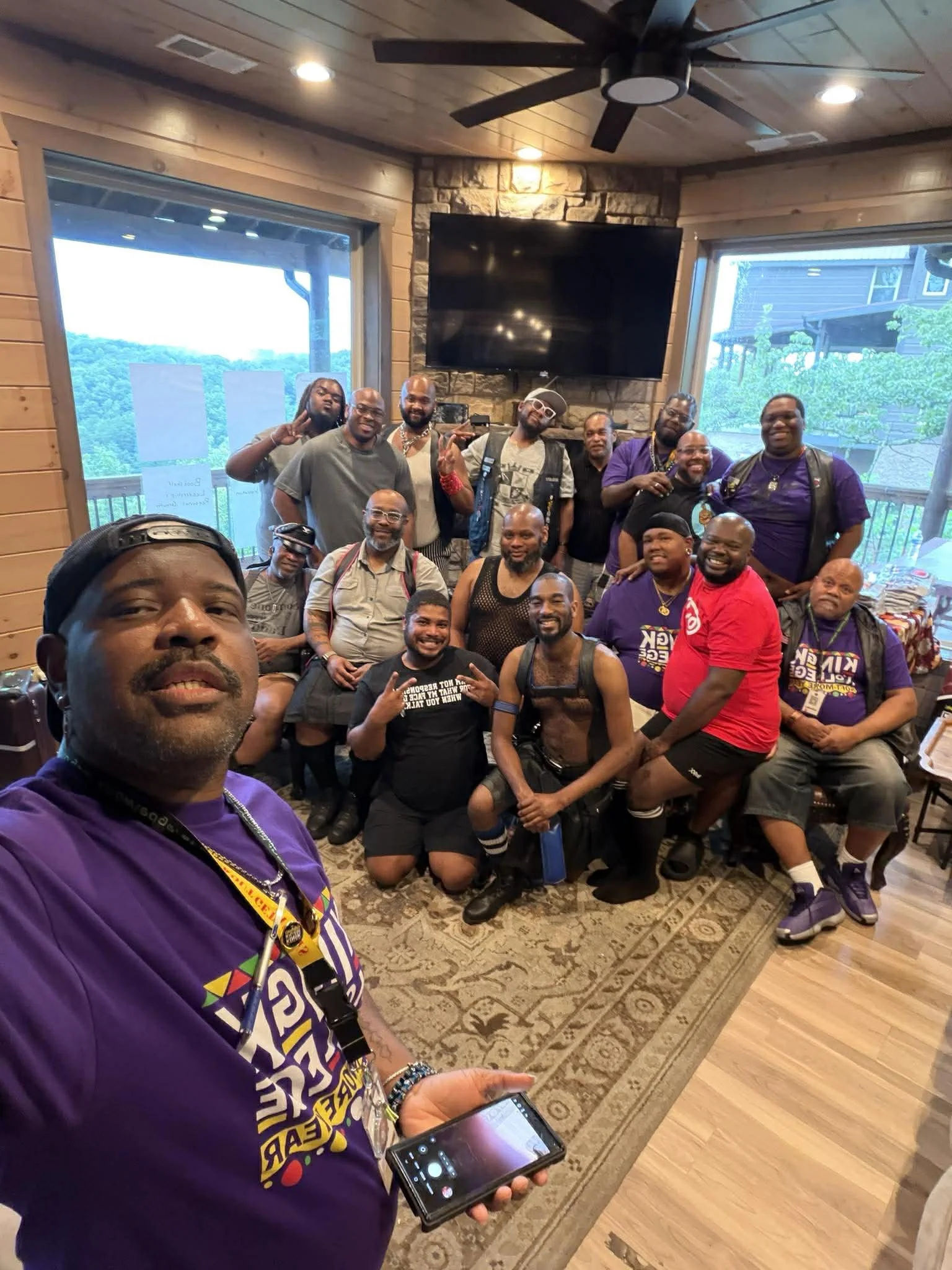 A group of diverse men in casual and athletic clothing gathered indoors in front of large windows with scenic mountain views, posing for a group photo at a social event or gathering.