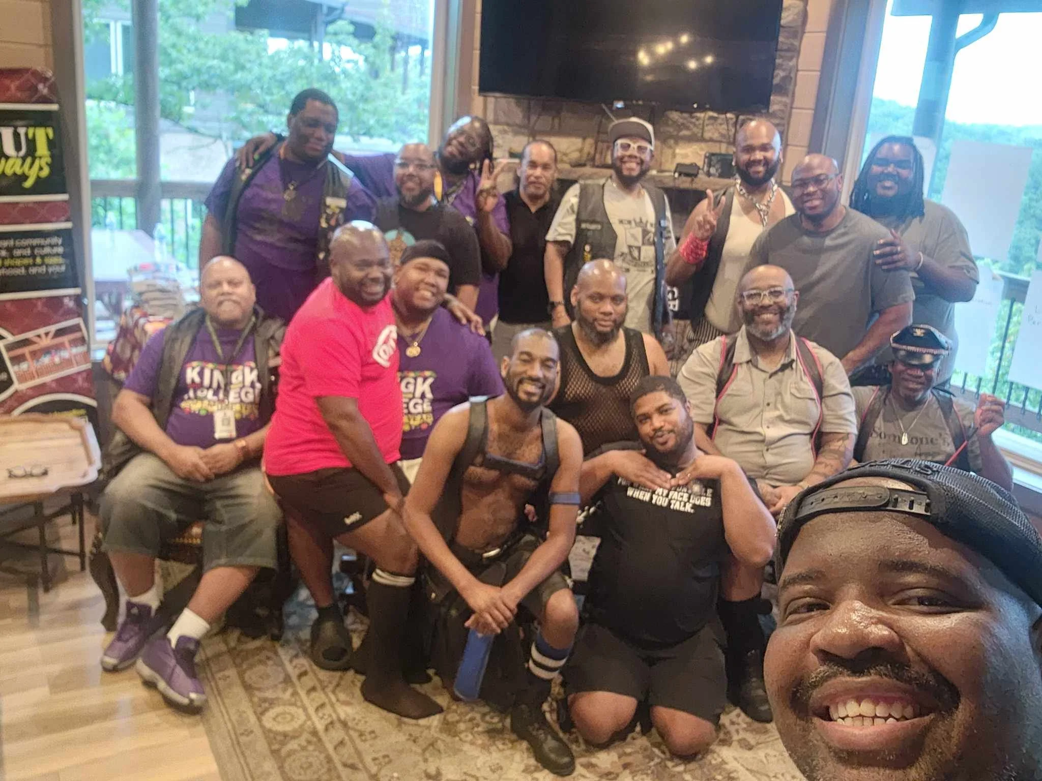 Group of people posing together indoors, in front of a large TV and stone fireplace, with some smiling and making peace signs.