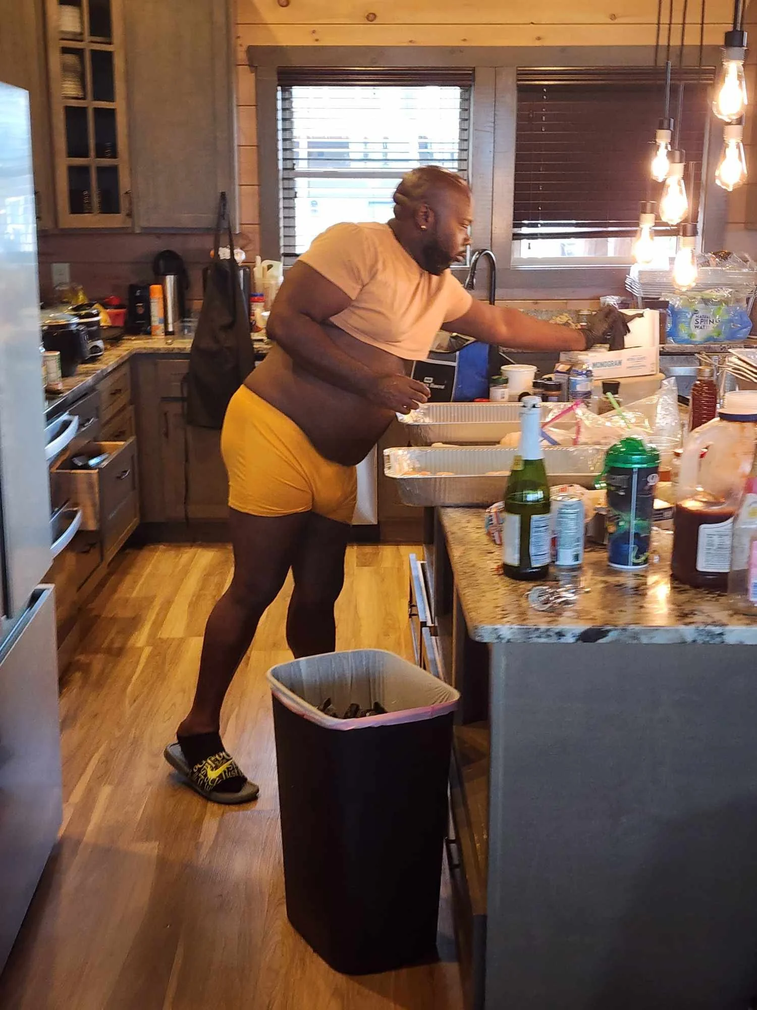 A man with dyed blonde hair and a beard, wearing a tan T-shirt, yellow shorts, and slippers, is cleaning a kitchen counter. The kitchen has wooden cabinets and a granite countertop. There are various items on the counter, including bottles, cups, and