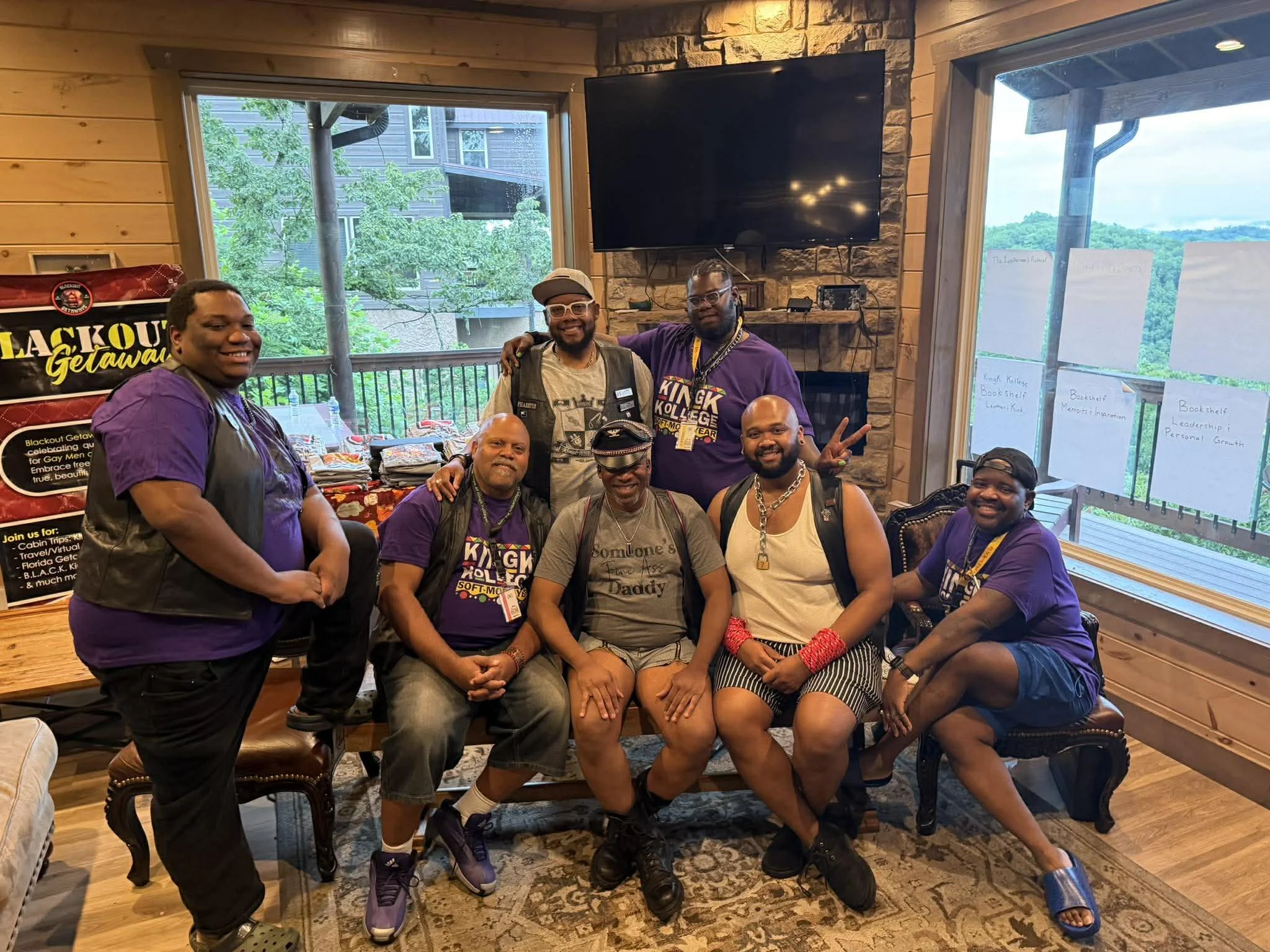 Group of eight diverse men gathered together indoors, smiling and posing for the photo. They are in a cozy, wood-paneled room with large windows showing a green outdoor view, and a stone fireplace with a mounted flat-screen TV. Some men are seated, a