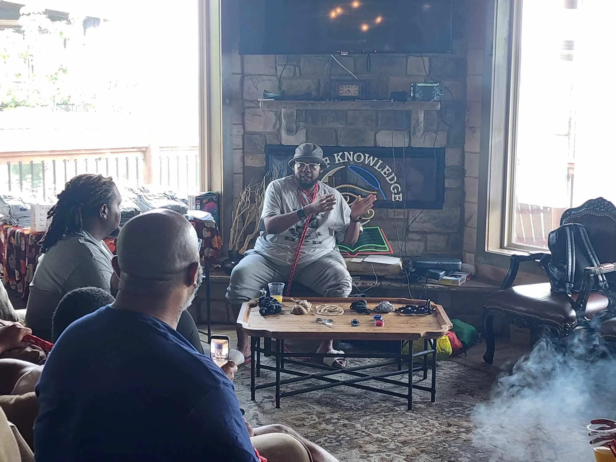 A man wearing sunglasses, a hat, and casual clothing is sitting on a chair in front of a brick fireplace, speaking to an audience gathered in a living room. The audience is sitting on couches and chairs, listening attentively. A coffee table with var