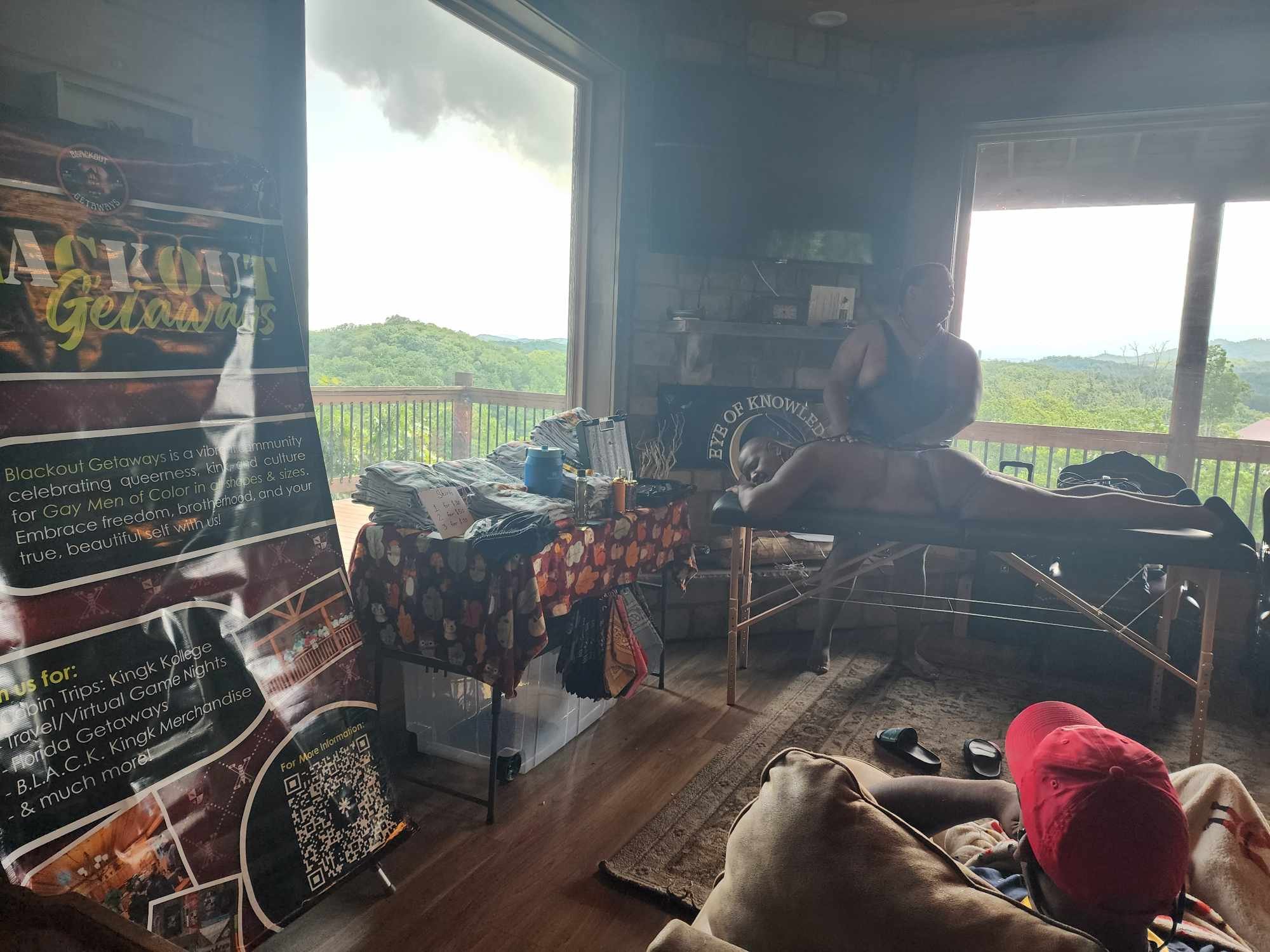 Interior of a room with a large window showing a green landscape outside. There is a person lying on a table, a table with folded t-shirts, a pink container, and multiple pairs of shoes and clothing on the floor. A poster promoting Blackout Getaways 