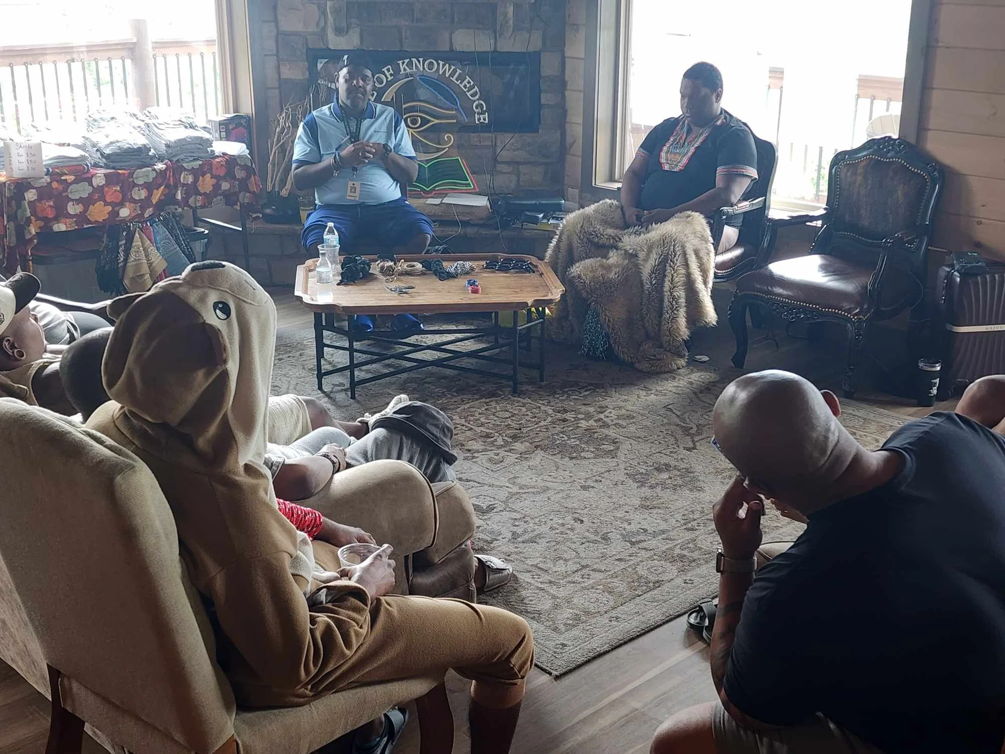 A group of people seated in a cozy room, with two men sitting in chairs facing each other, one wearing a traditional shirt, the other in a casual outfit; a woman in a bear hoodie is visible in the foreground. The room has a stone fireplace with a scr