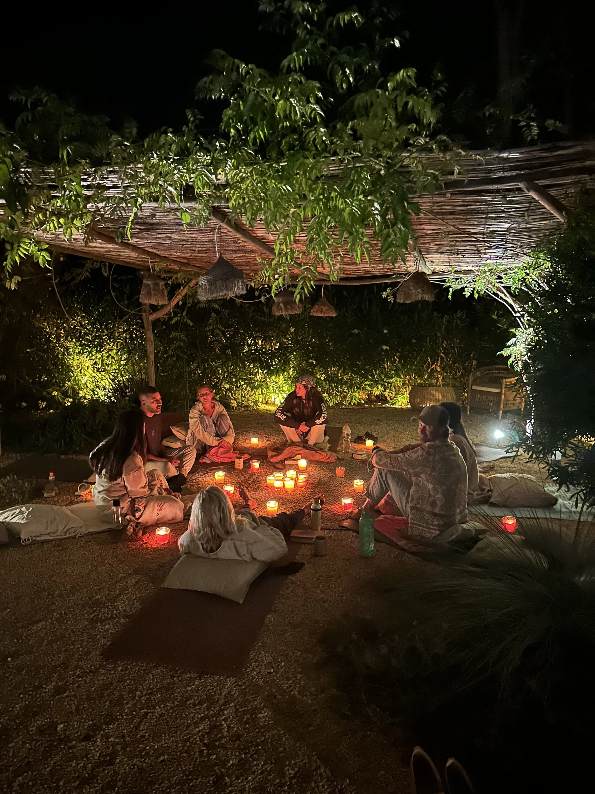 People sitting in a circle on the ground around candles at night outdoors, under a thatched roof, with trees and greenery around.