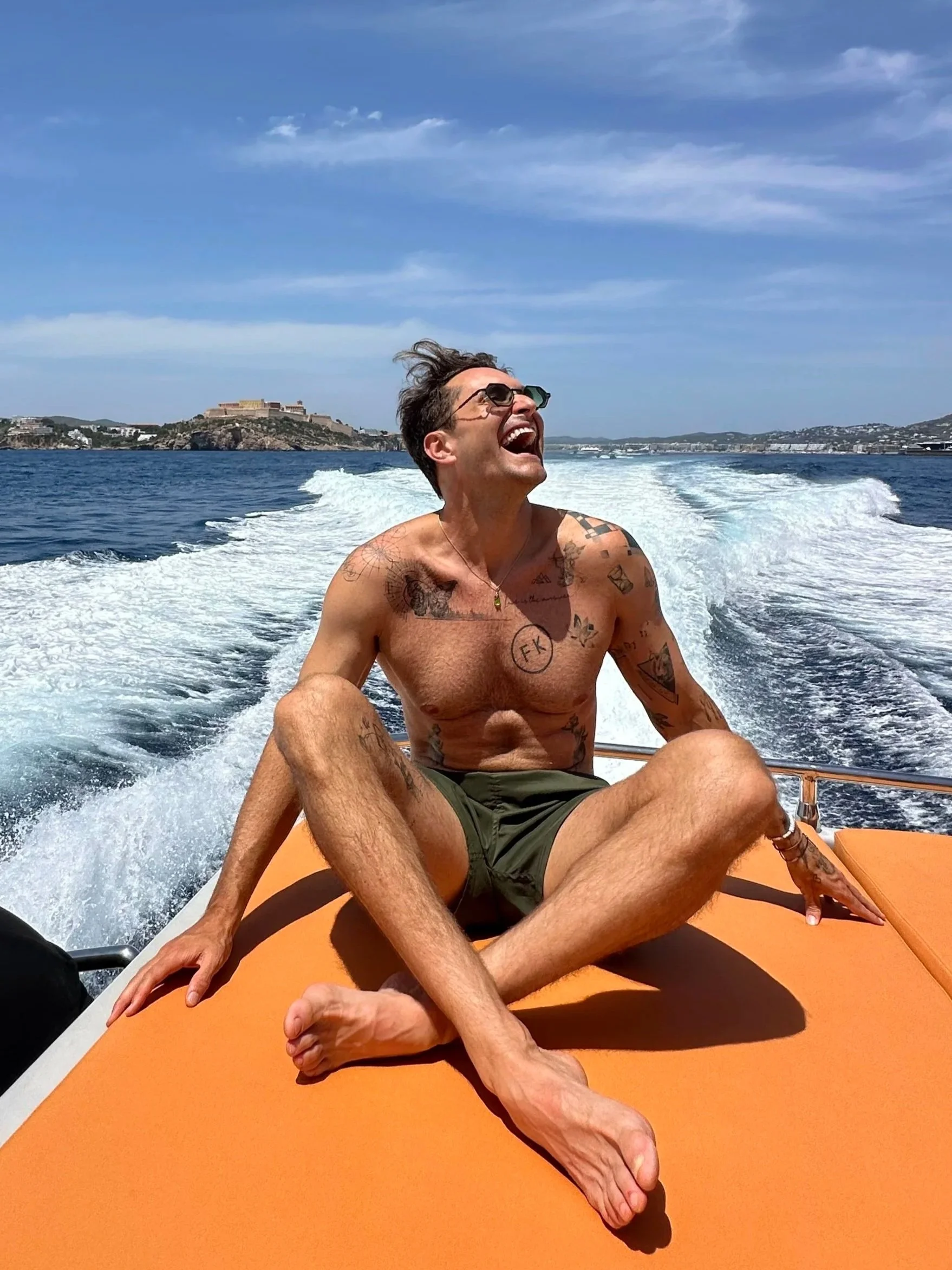 A shirtless man with tattoos smiling and laughing on a boat during daytime, with water and distant shoreline in the background.