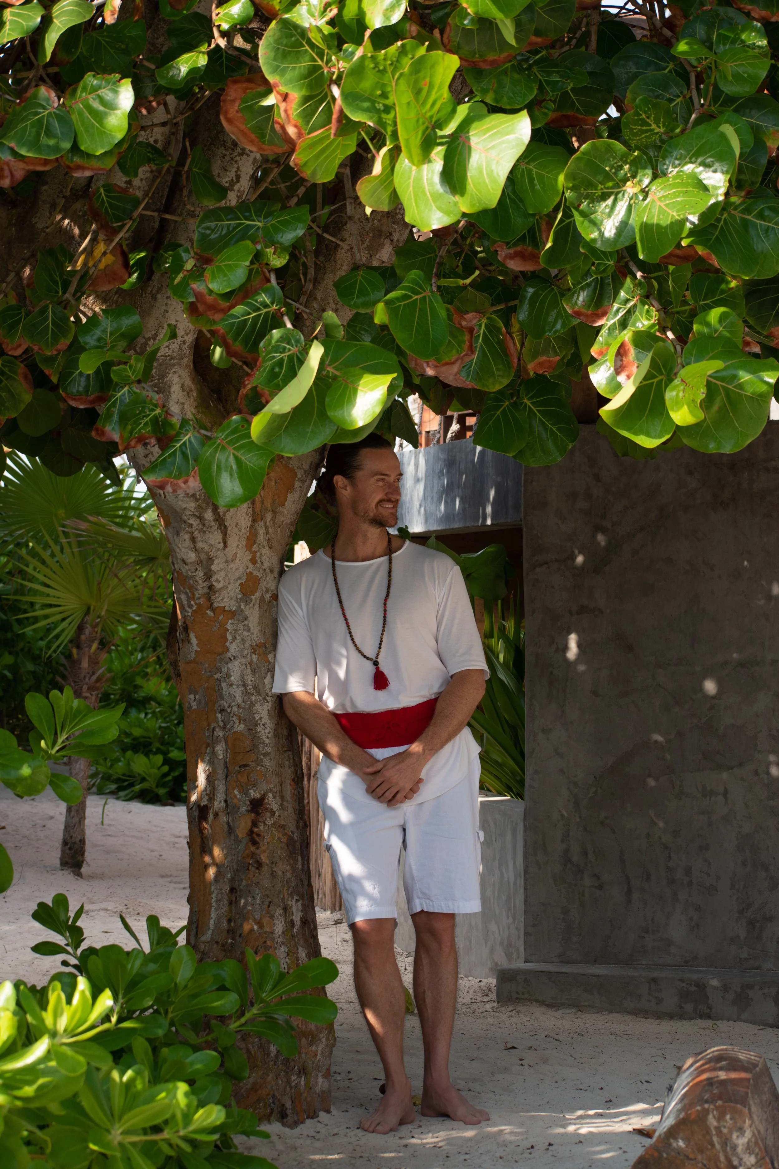 A man in white attire leaning against a tree on a beach, surrounded by lush green foliage, with sandy ground and a concrete structure in the background.