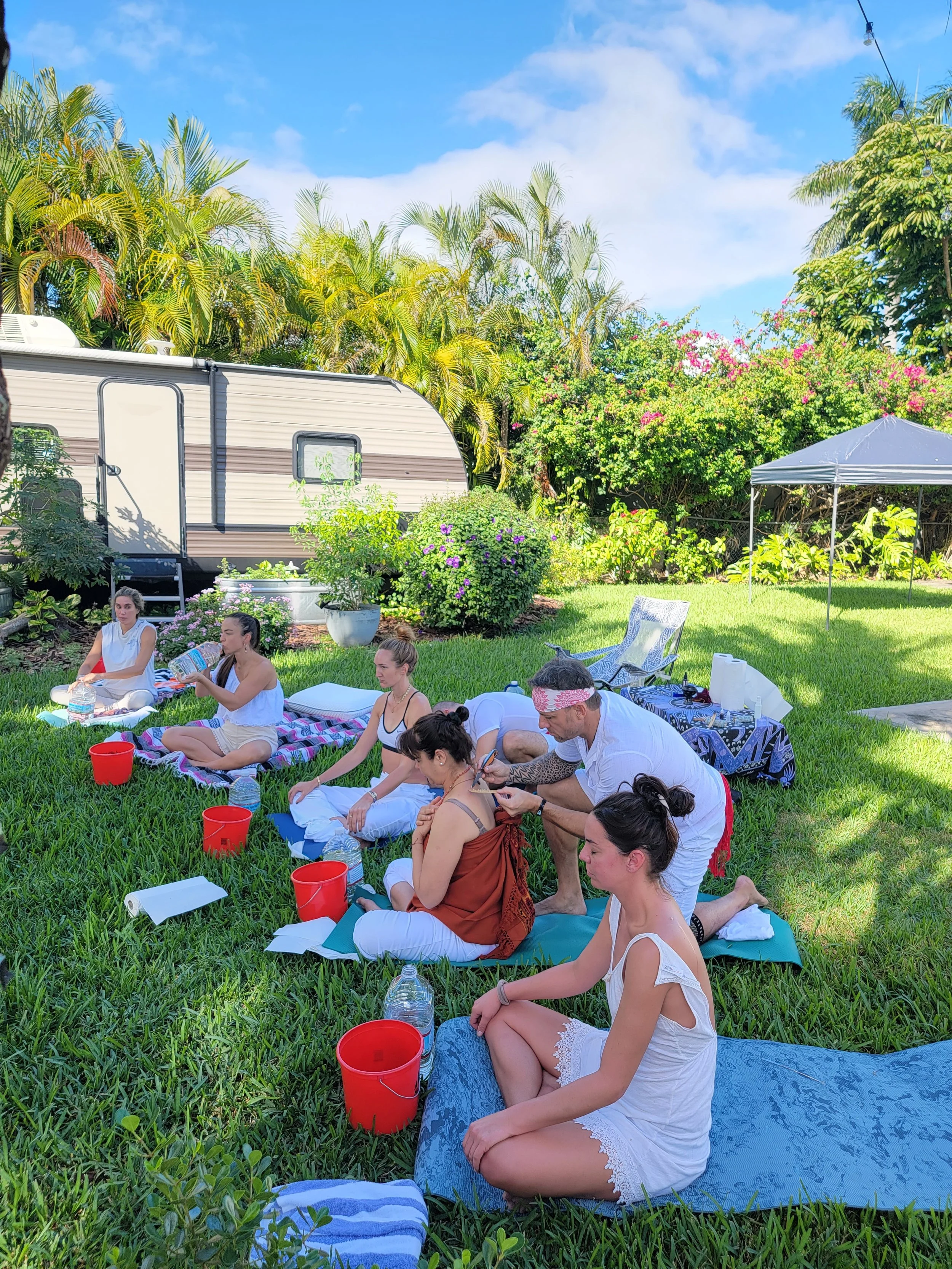 Group of people participating in a healing or meditation session outdoors on a grassy area, with a caravan, trees, and a blue sky in the background.