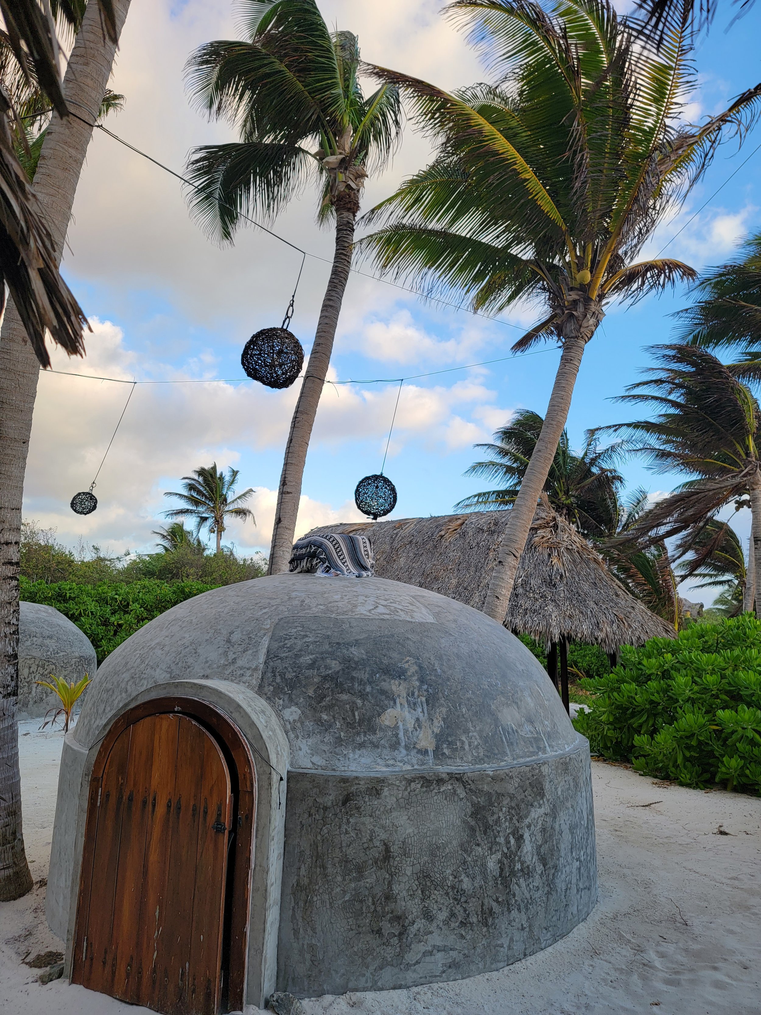 Beach scene with palm trees, spherical lanterns hanging, a small concrete dome-shaped hut with a wooden door, and thatched roof hut in background.