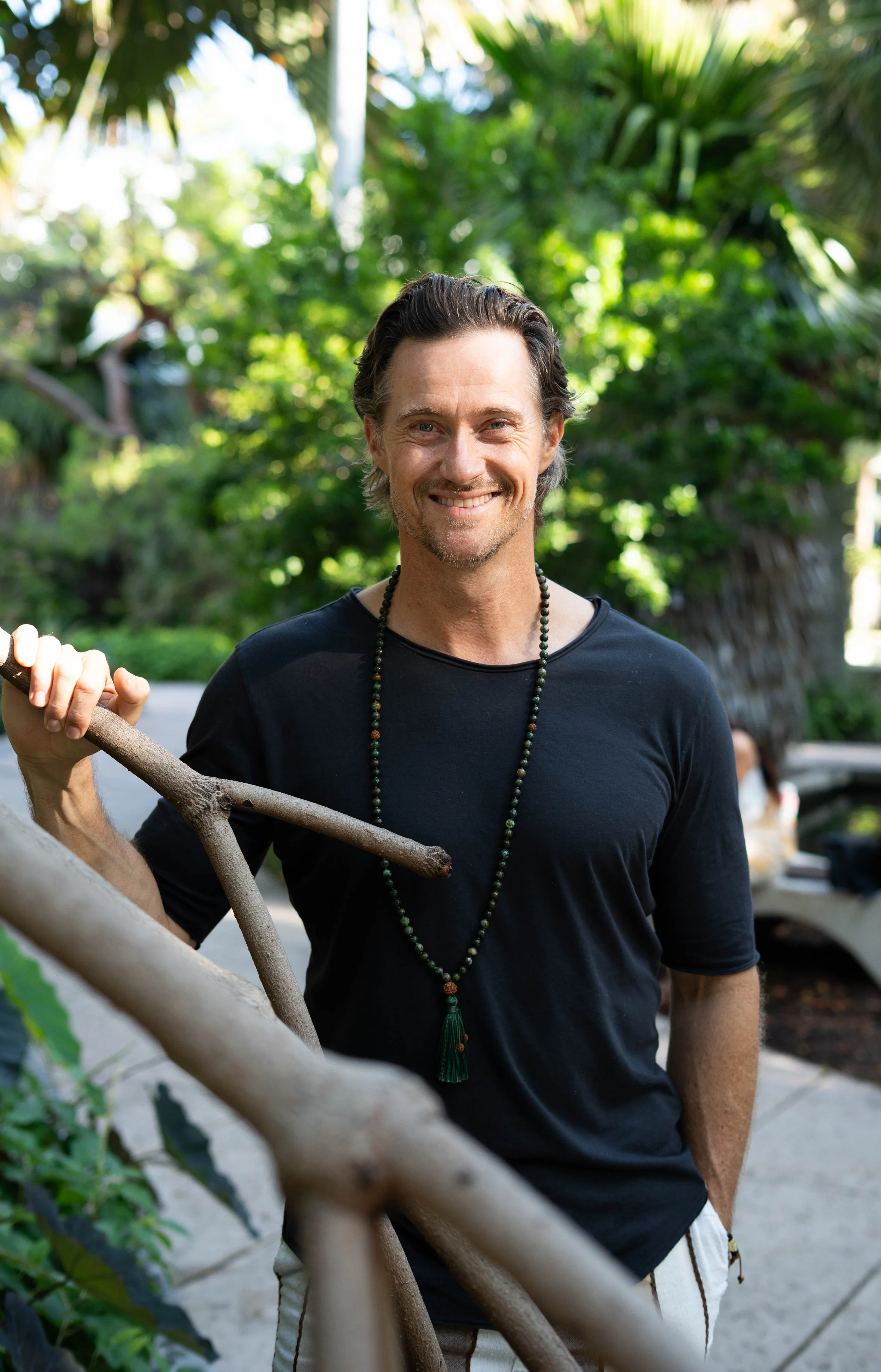 A smiling man with dark hair and a beard, wearing a black t-shirt and beaded necklace, standing outdoors in a lush green setting, holding a branch with his left hand.