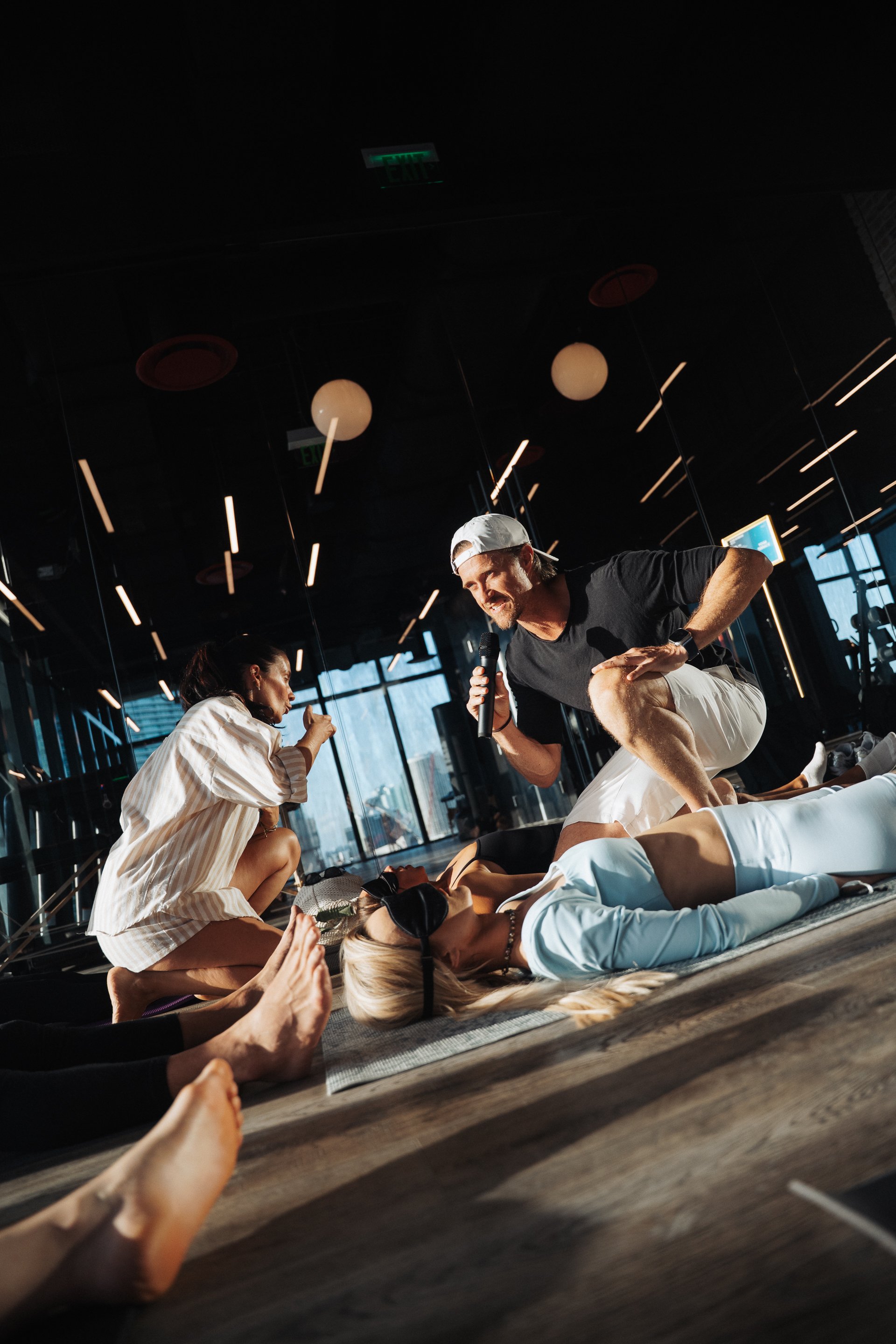 A man with a microphone speaking to a group of people lying on mats, some wearing eye masks, in a dimly lit room with large windows.