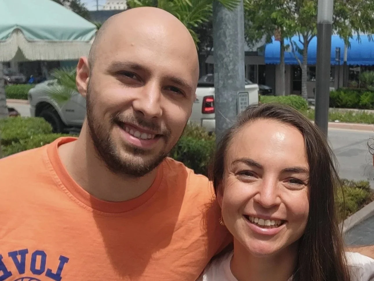 Two smiling people, a man with a beard in an orange T-shirt and a woman with long brown hair, standing outdoors in a parking lot with green shrubs, trees, and parked cars in the background.