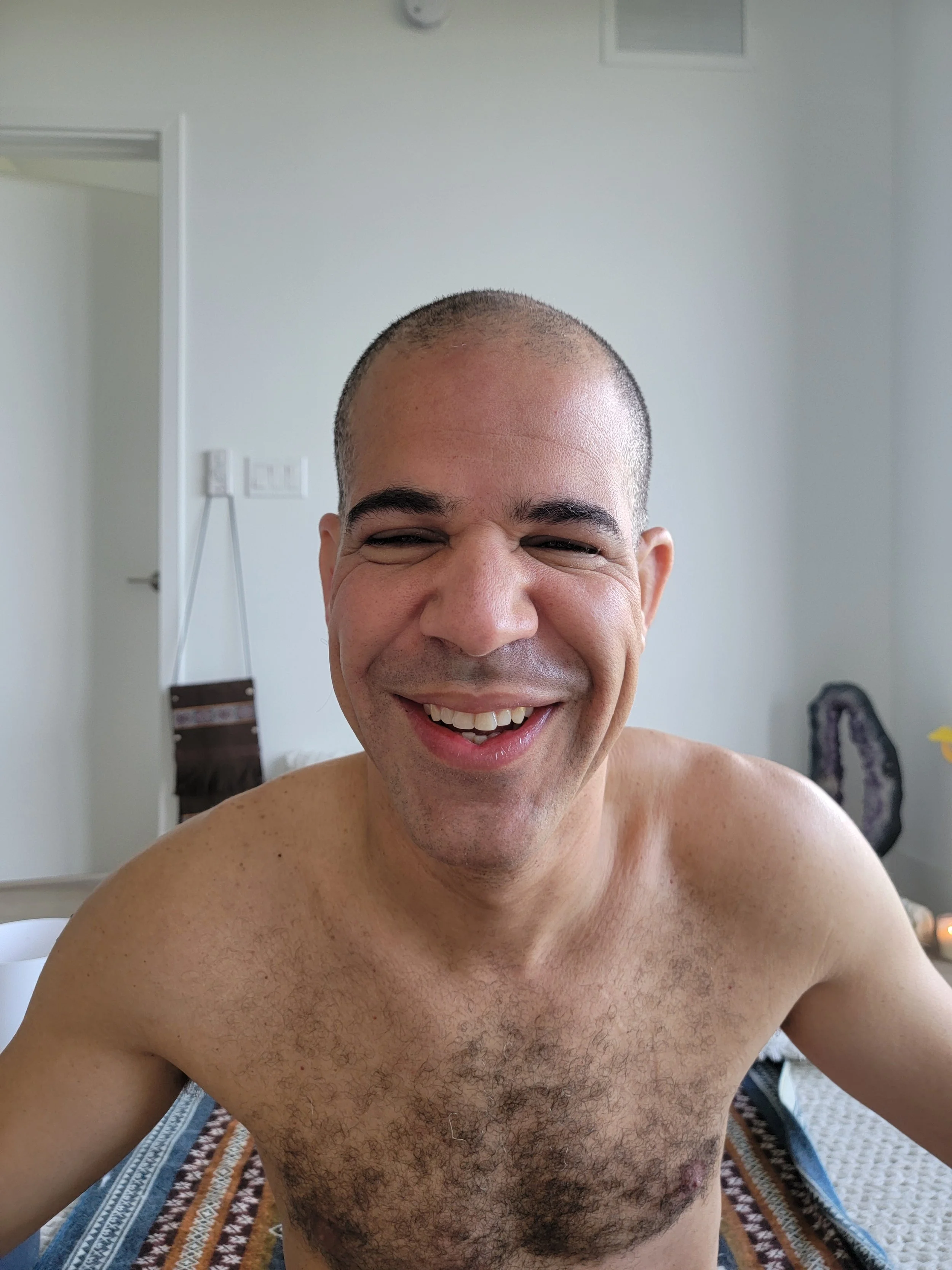 A shirtless man with short hair smiling and laughing in a room with white walls and minimal decor.