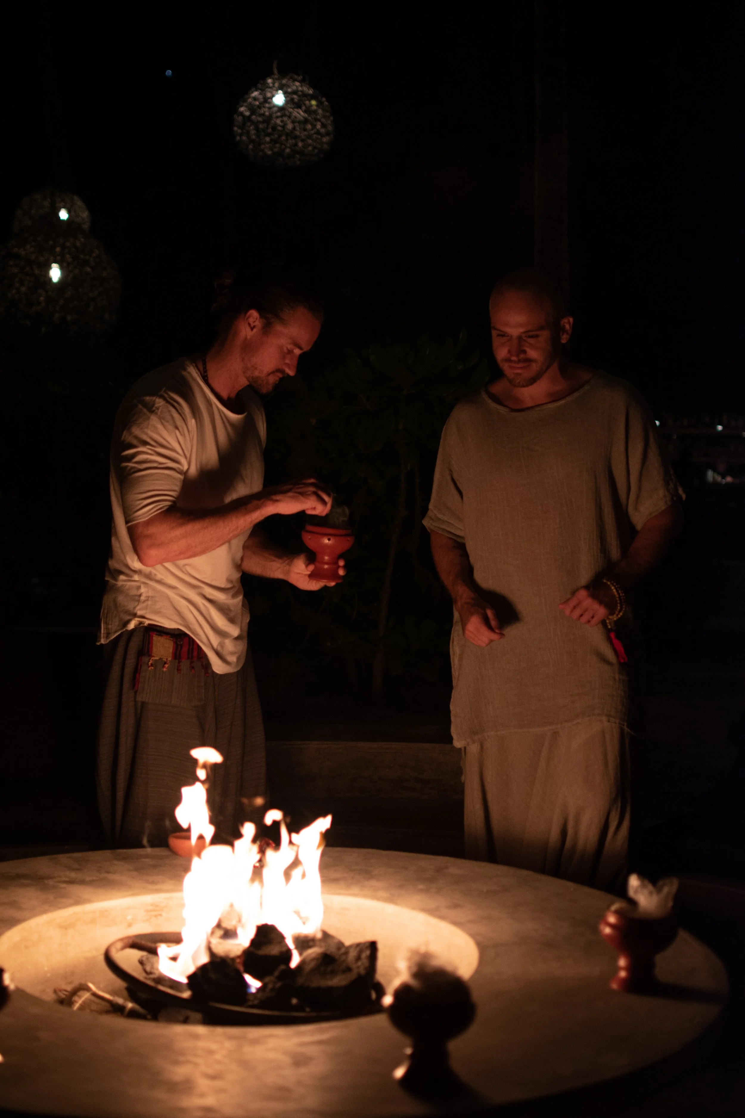 Two men standing around a fire pit at night, one holding a small object, the other with a phone, with hanging lights in the background.
