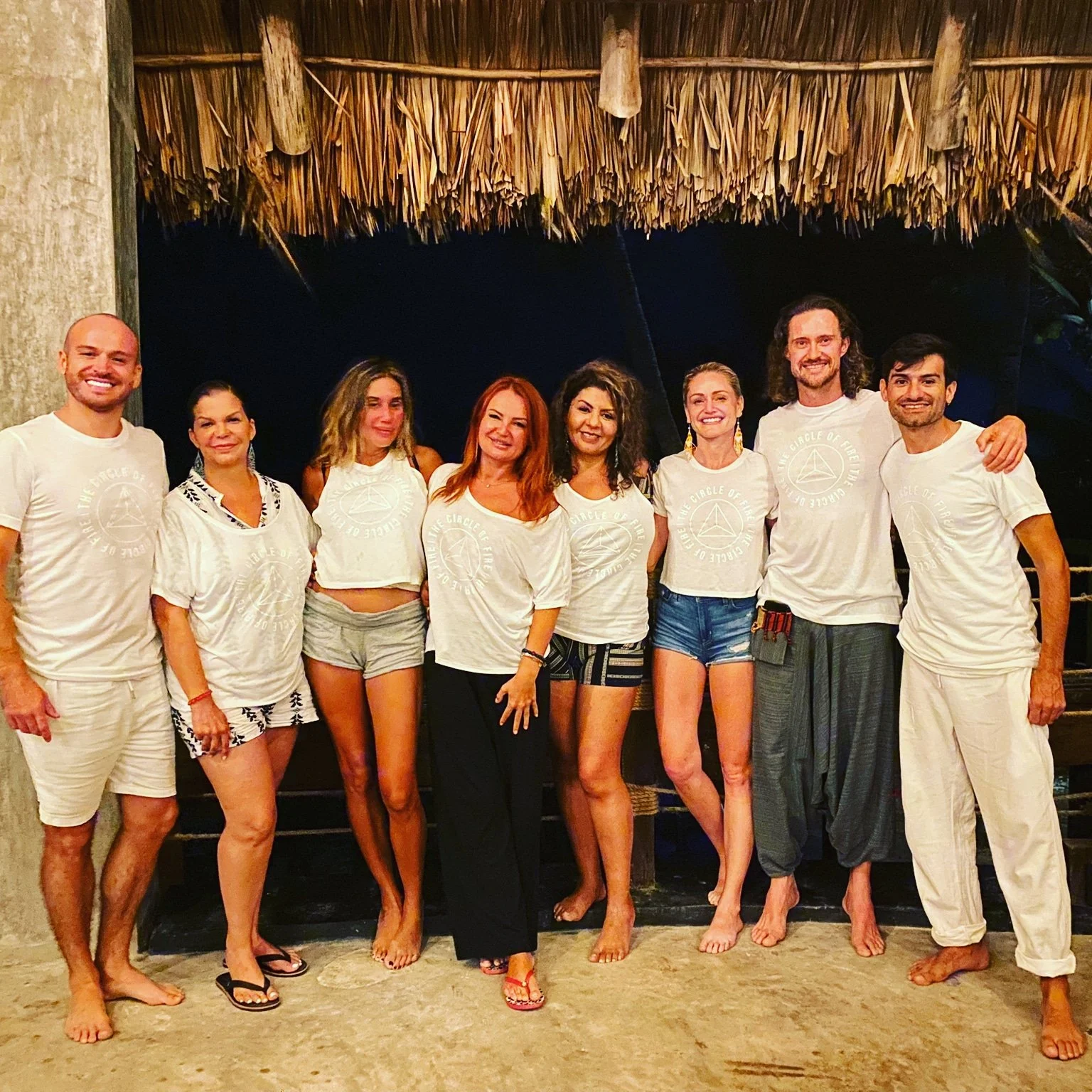 Group of people standing together at a tropical or beachside setting, wearing casual summer clothing, several in matching white T-shirts with a circular logo, smiling for the camera.