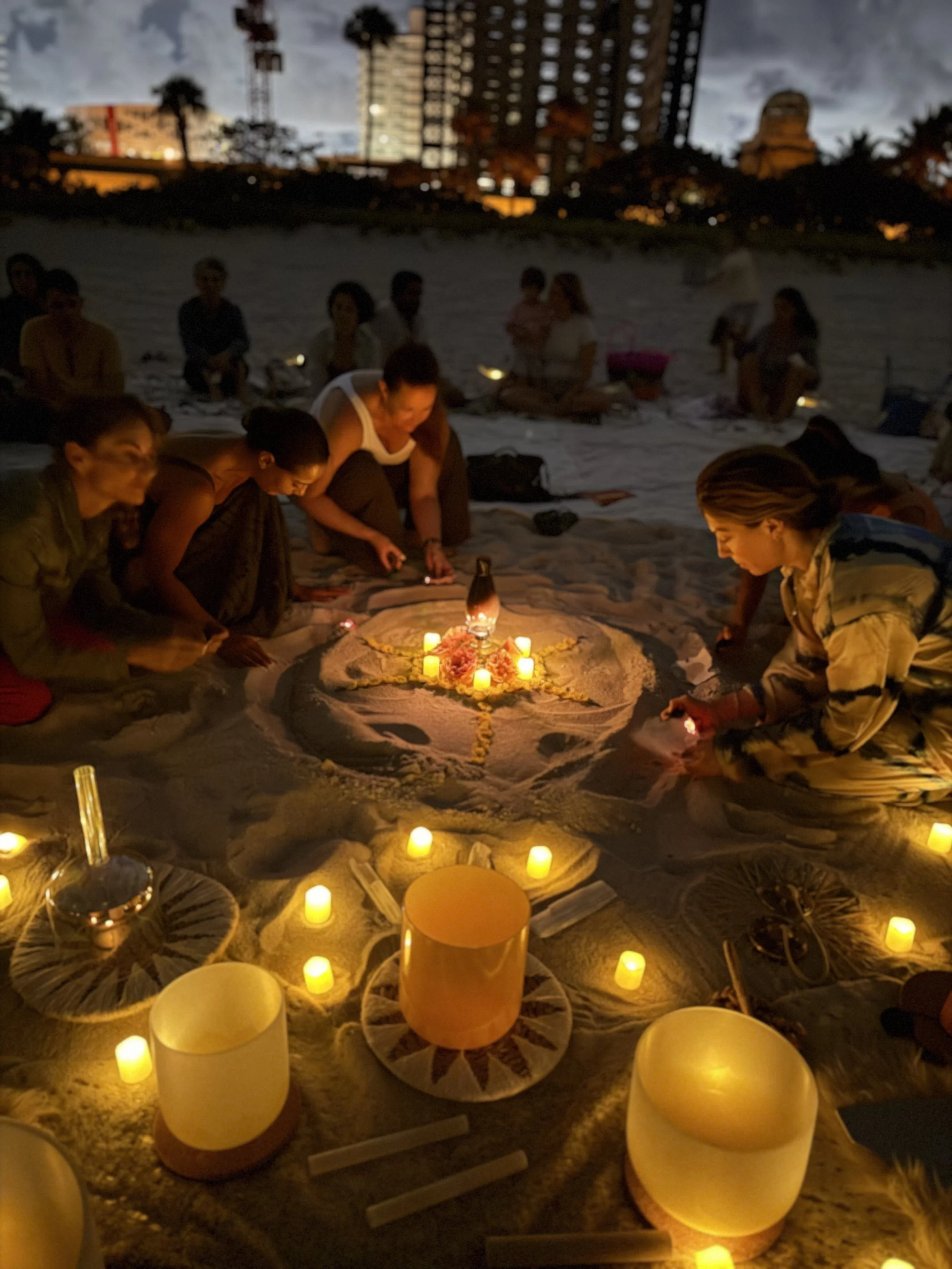 Group of people gathered on a beach during fullmoon, participating in a candlelit ceremony or meditation with candles and decorative objects in the sand, with buildings and trees in the background.