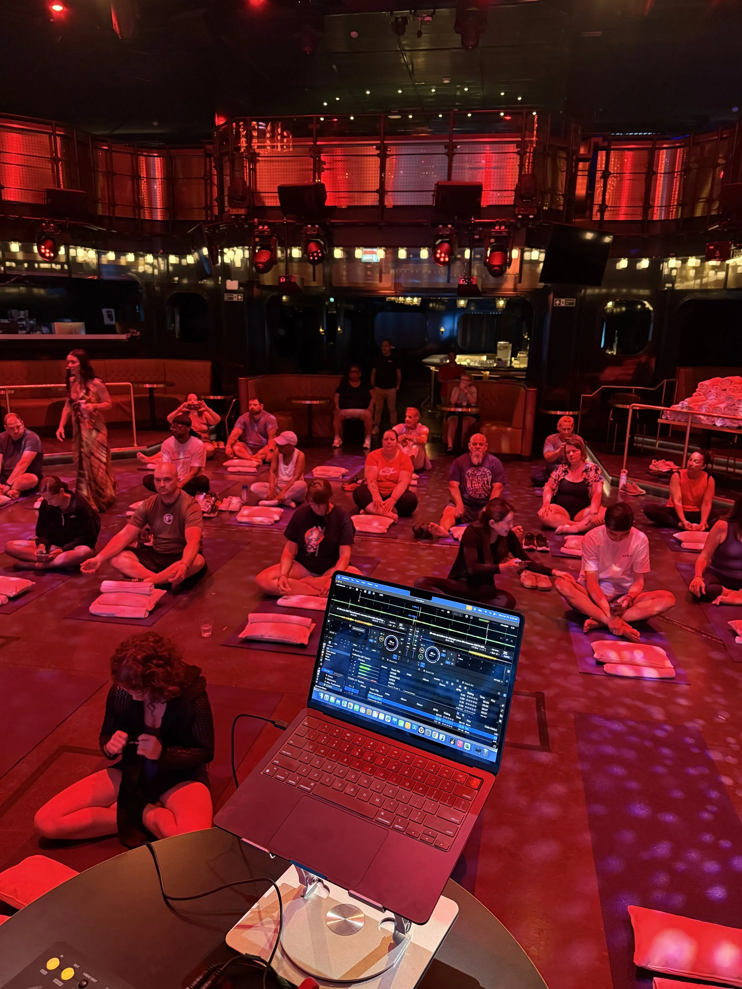 People seated cross-legged on the floor with cushions during a breathwork session in a dimly lit, red-hued room with stage lights and a DJ setup in the foreground.