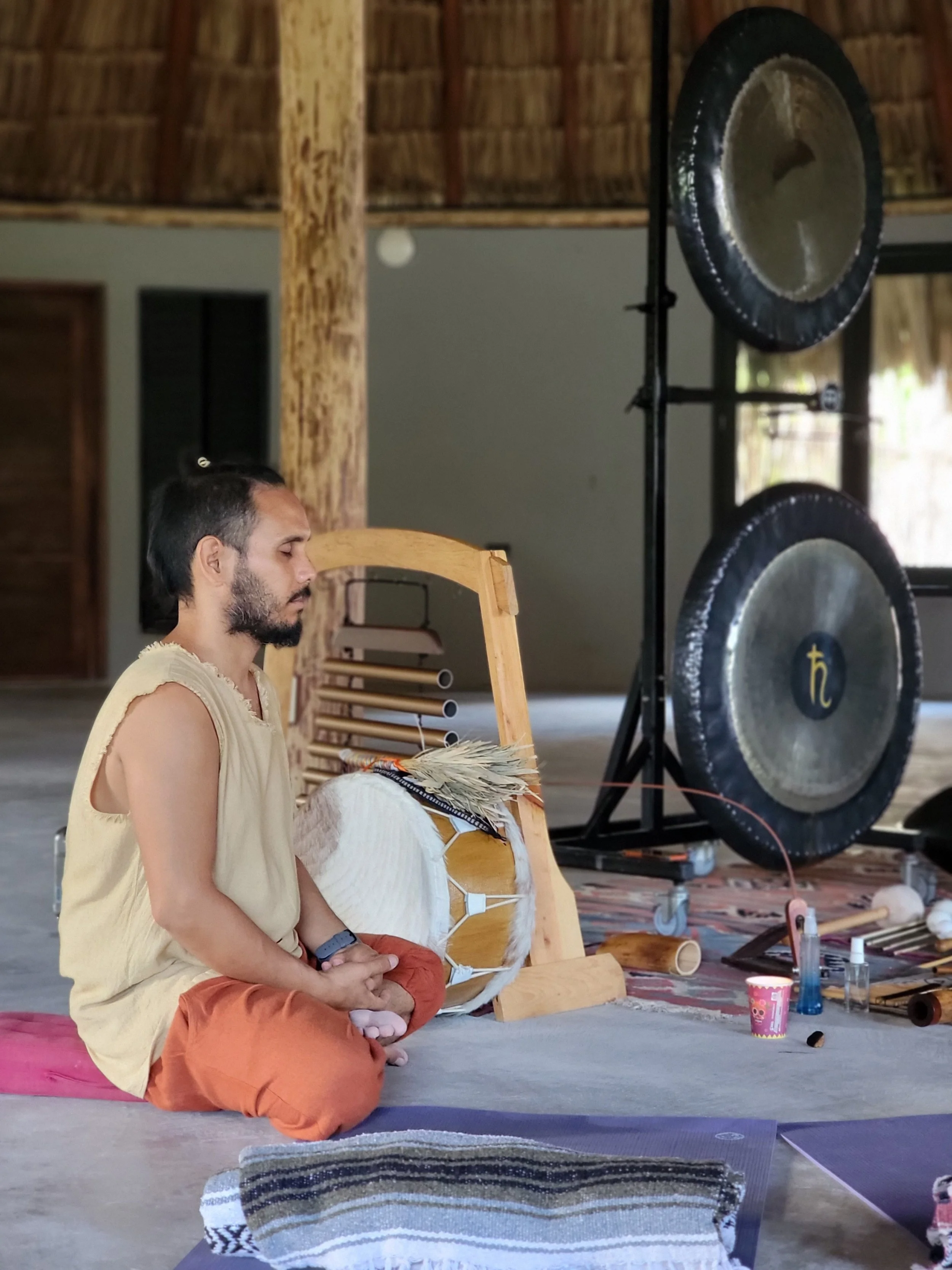 A man with closed eyes and crossed legs in a cross-legged sitting position, practicing meditation or prayer, surrounded by musical instruments including gongs, drums, and flutes in a peaceful indoor setting with wooden elements.