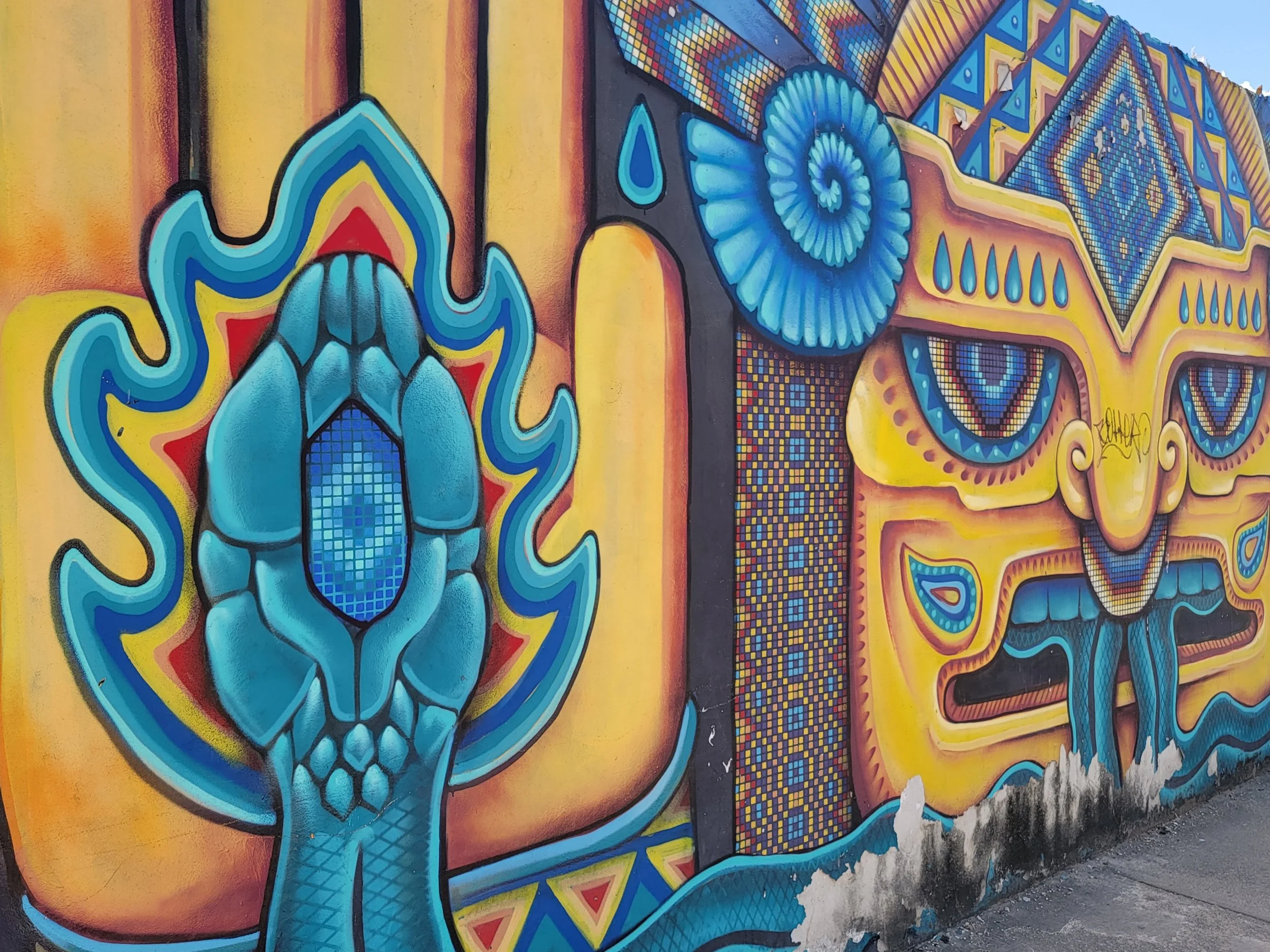Colorful mural of a stylized face with intricate patterns, a blue eye motif, and vibrant geometric designs in yellow, blue, and red.