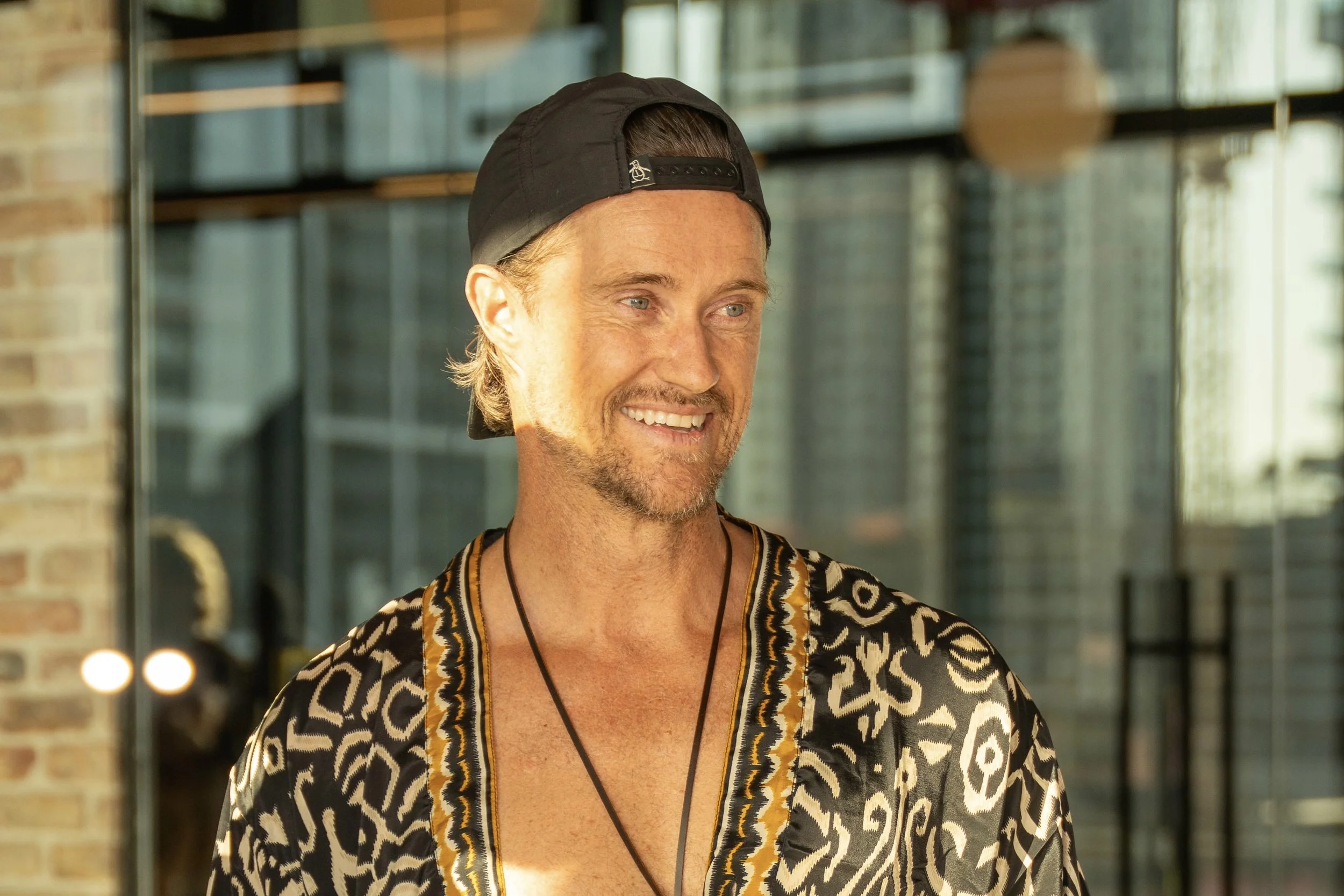 A smiling man with a beard and mustache, wearing a backwards black cap and a patterned shirt, standing indoors near a large window with a cityscape in the background.