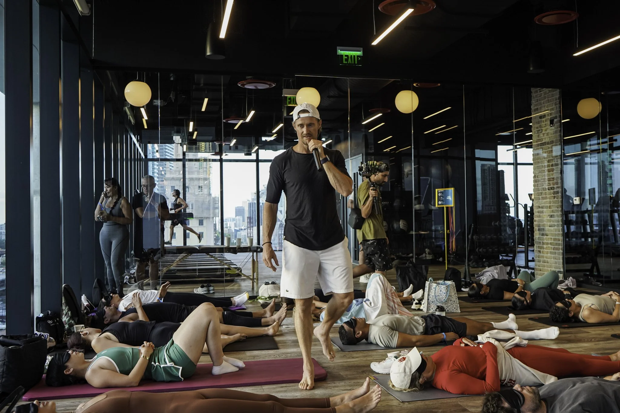 Indoor yoga class with people lying on mats and a man standing in the center holding a microphone, with large windows and city skyline in the background.