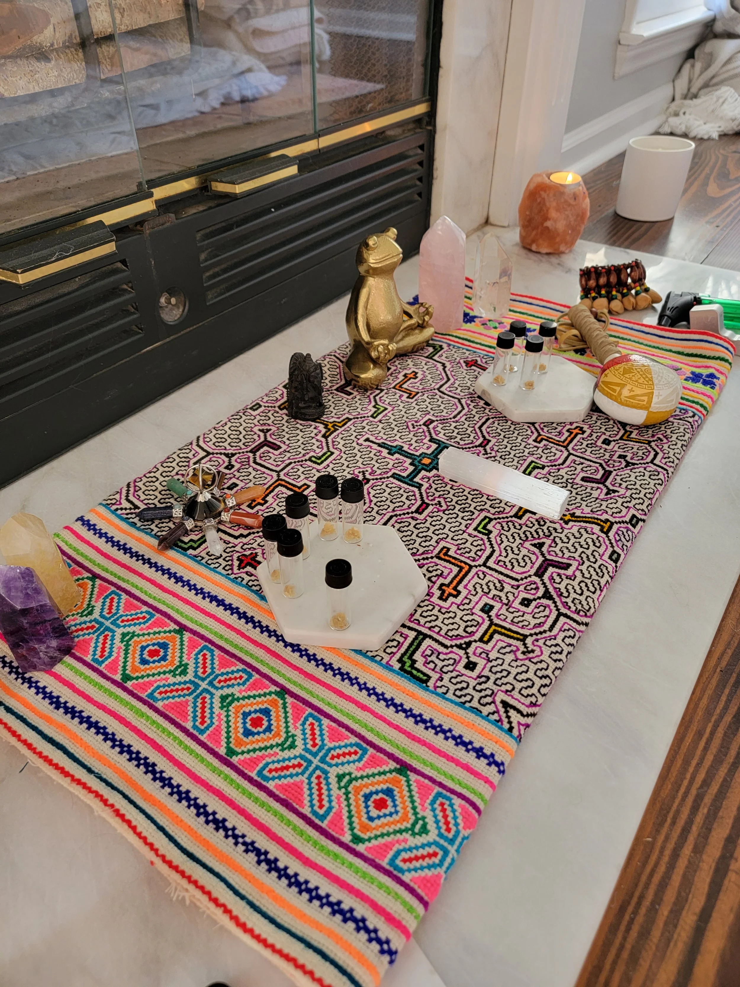 A spiritual or ritual setup on a colorful woven cloth includes crystals, small bottles, a golden frog figurine, a black sculpture, a salt lamp, a candle, and various small objects, placed beside a fireplace.