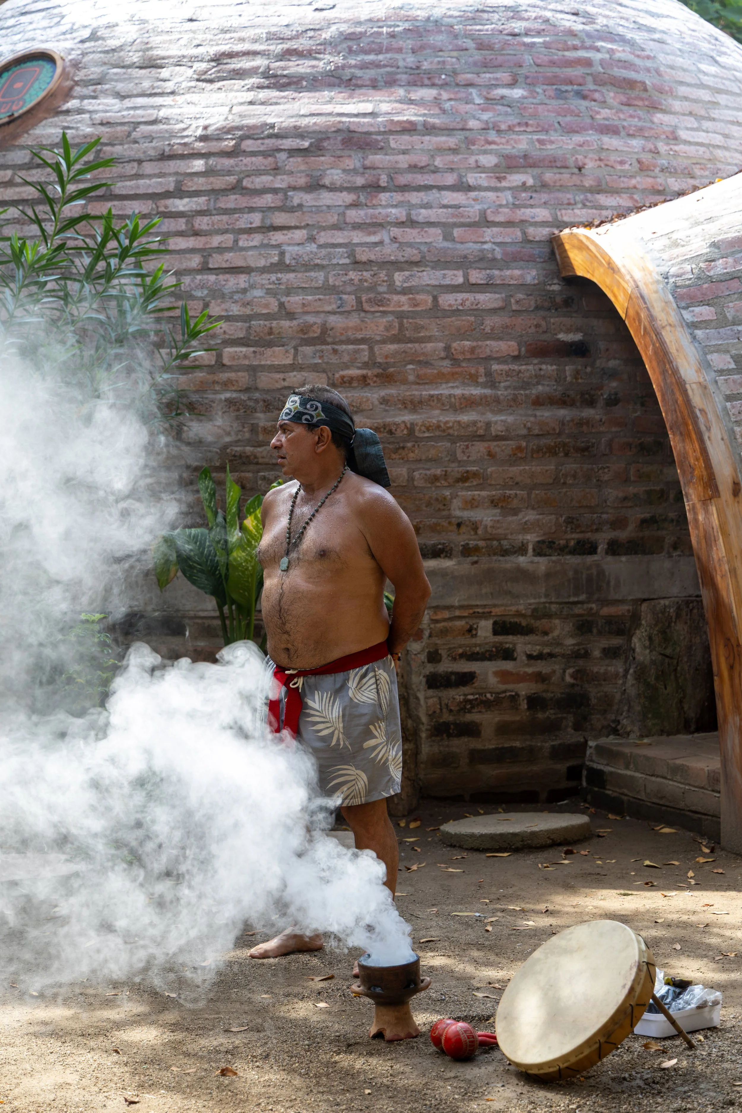 A shirtless man wearing a headband, beads, and patterned shorts stands outdoors near a smoking incense holder, with drums and a wooden structure nearby, against a brick wall background.
