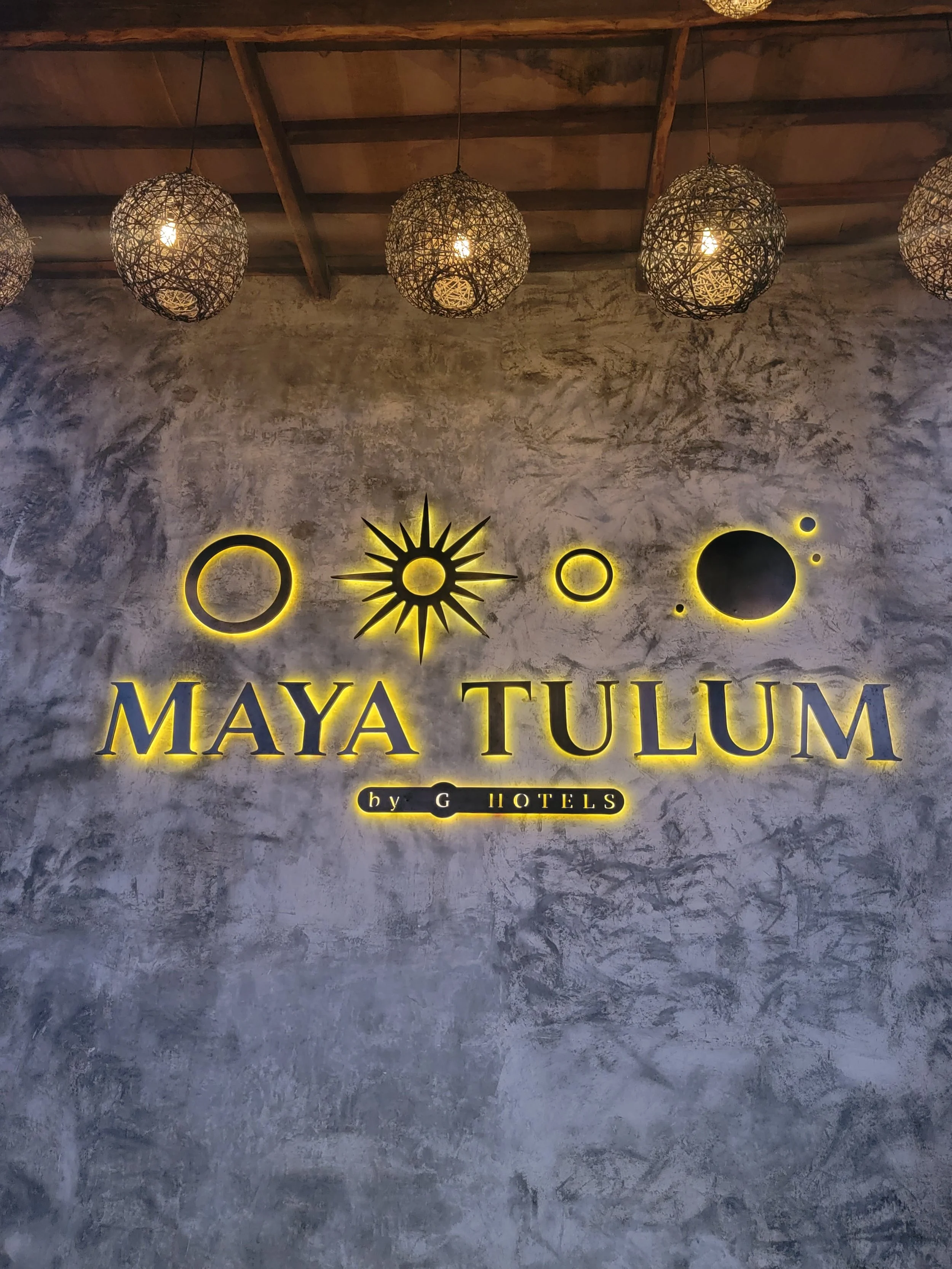 Neon sign on a textured concrete wall displaying the name 'Maya Tulum' with moon and sun graphics above it and 'by G Hotels' below, illuminated with yellow lights. Overhead, woven pendant lights hang from a wooden ceiling.