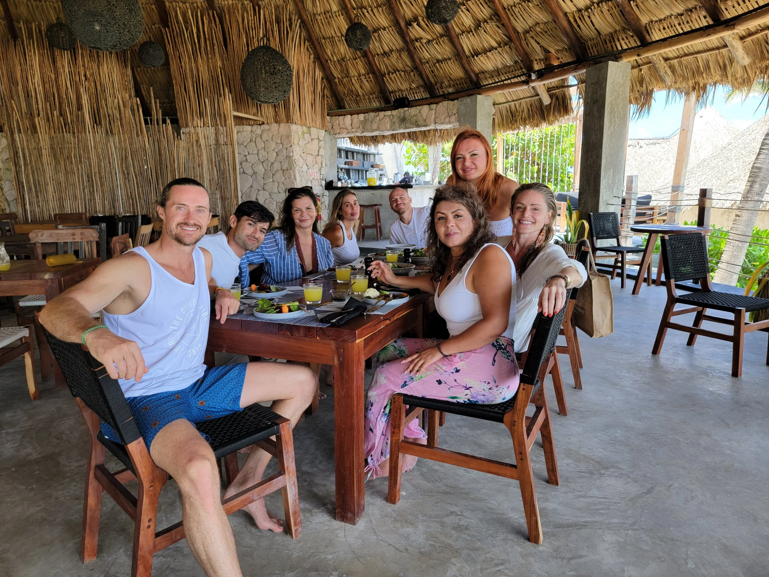 A group of nine people sitting and standing around a wooden dining table with food and drinks at a beachside restaurant with a thatched roof and open sides.