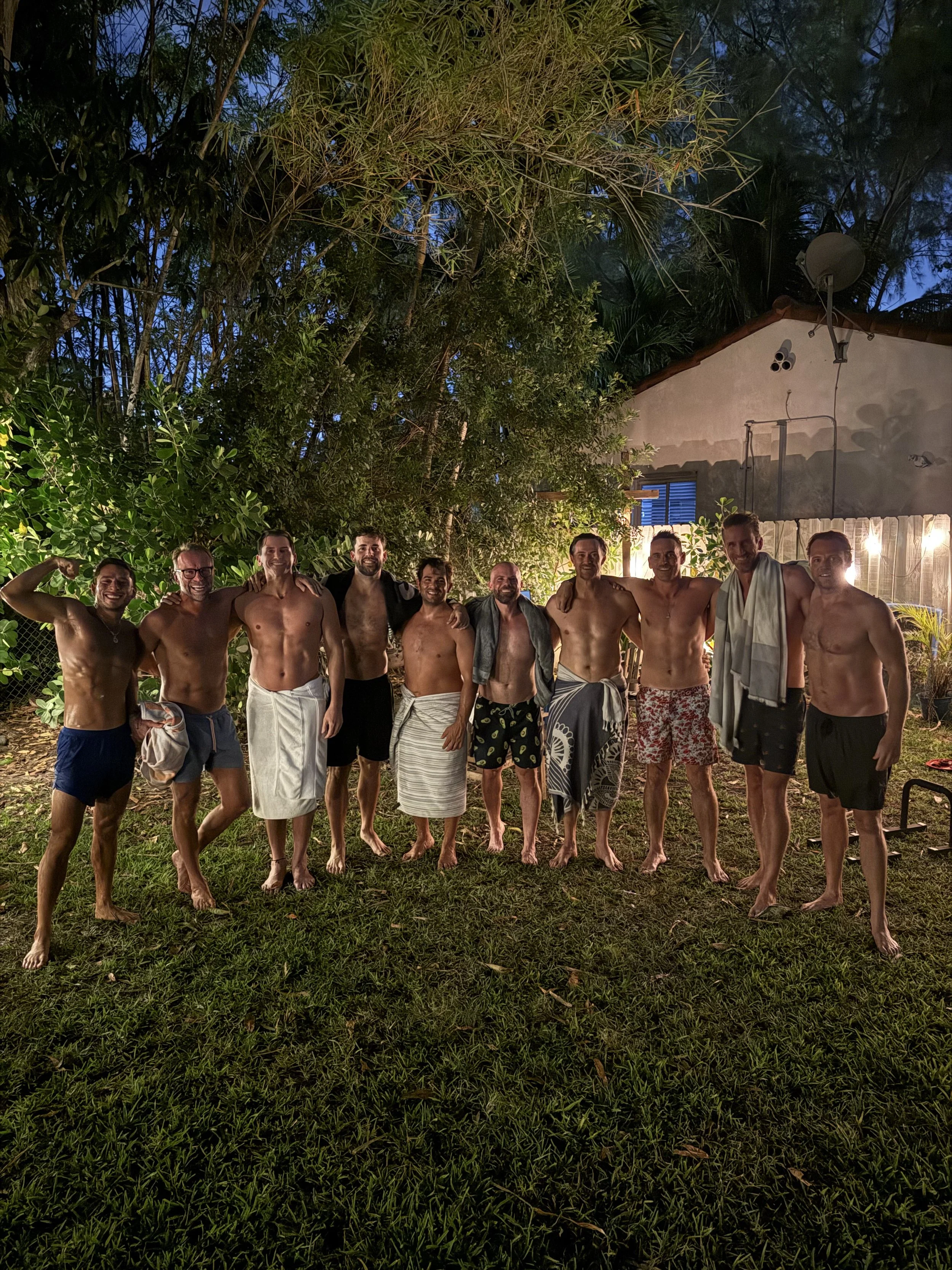 Group of ten men standing outdoors at night, some with towels, in front of a house with string lights, surrounded by trees and bushes.